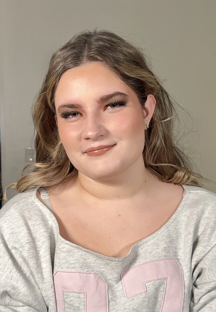 A woman with light brown, wavy hair styled in loose waves, wearing makeup including mascara, eyeliner, eyeshadow, blush, and glossy lipstick. She is dressed in a light gray, off-the-shoulder top with pink embroidered lettering. She is smiling slightl