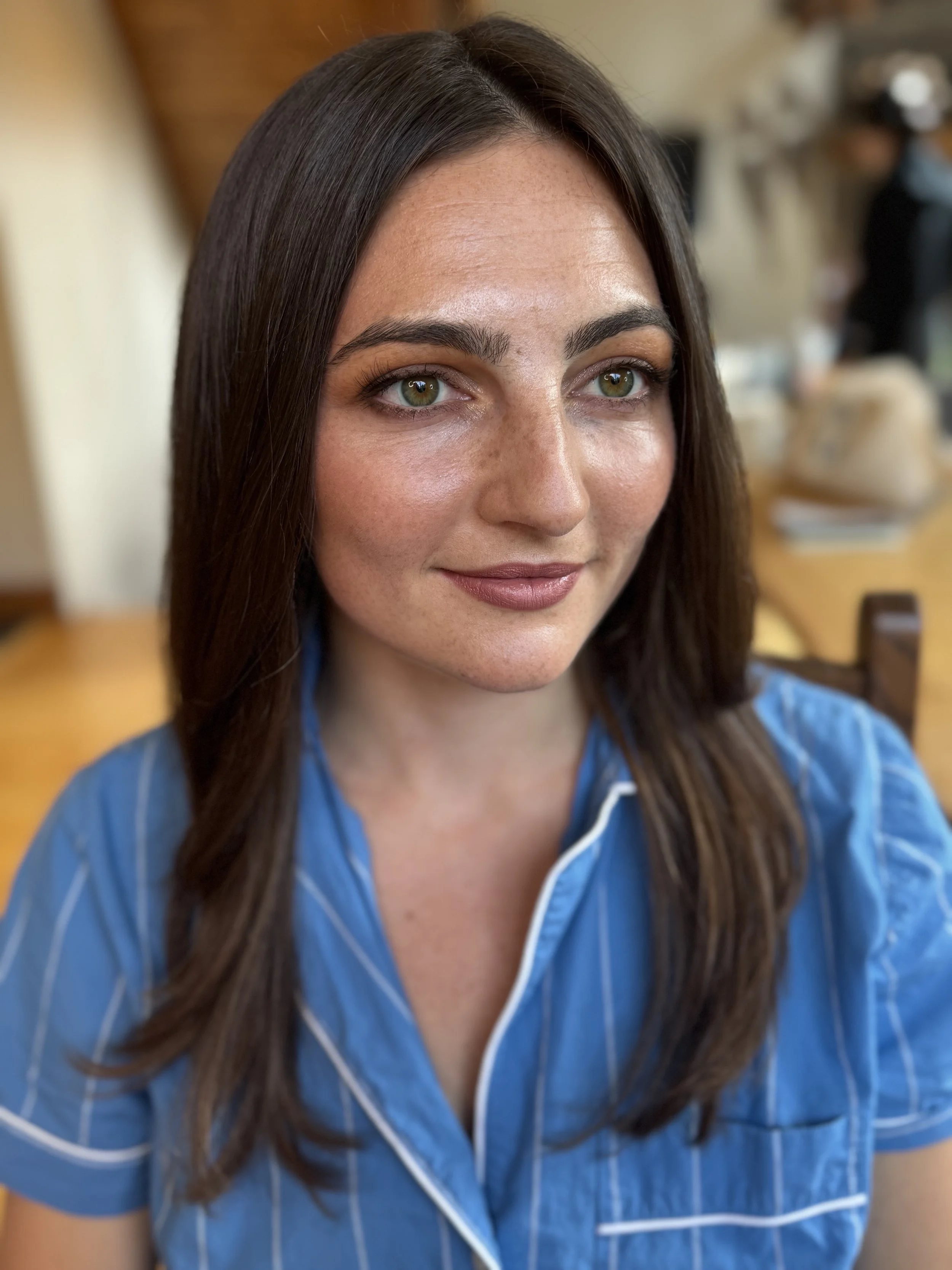 A young woman with long brown hair and green eyes, wearing a blue striped shirt, sitting indoors with a blurred background.