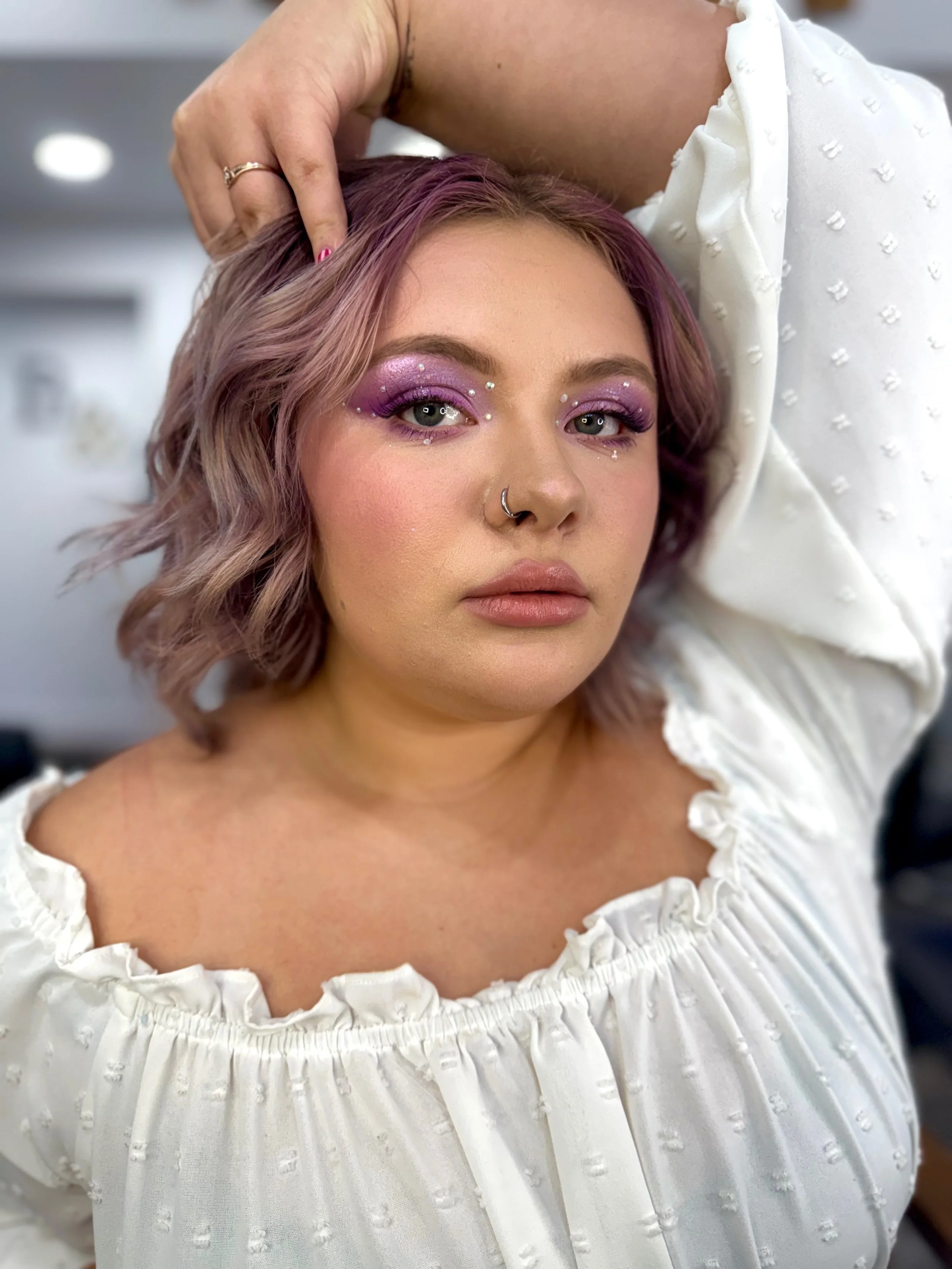 A woman with short, wavy purple hair and makeup with purple eyeshadow, rhinestone embellishments, and nude lipstick. She is wearing a white, off-the-shoulder blouse with ruffled edges and has a nose ring. An adult hand is gently touching her head.