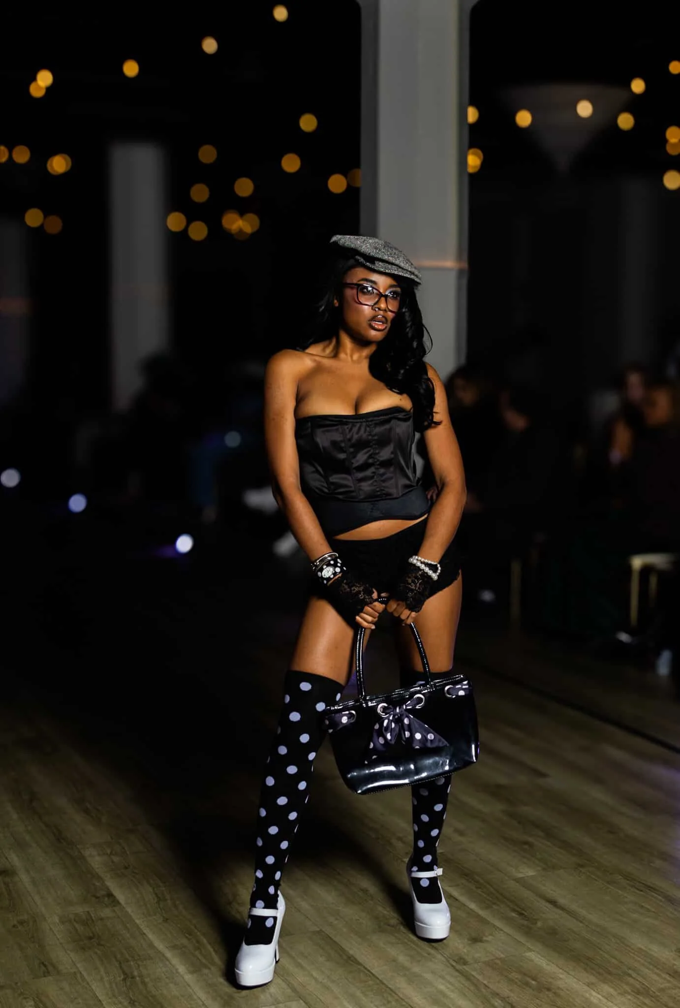 A woman with dark skin and black hair wearing glasses, a hat, a black strapless top, black shorts, polka dot thigh-high socks, and white platform shoes standing indoors with a dark background and bokeh lights.