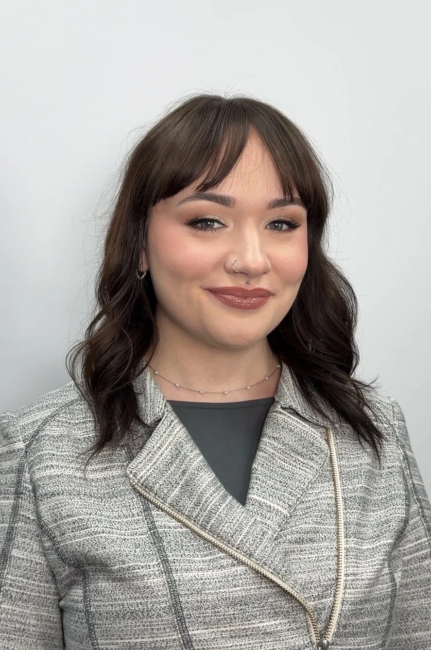 A woman with shoulder-length dark brown wavy hair, light skin, and blue eyes, wearing makeup, a nose ring, earrings, and a delicate necklace. She is dressed in a black top and a gray plaid jacket, posing against a plain gray background.
