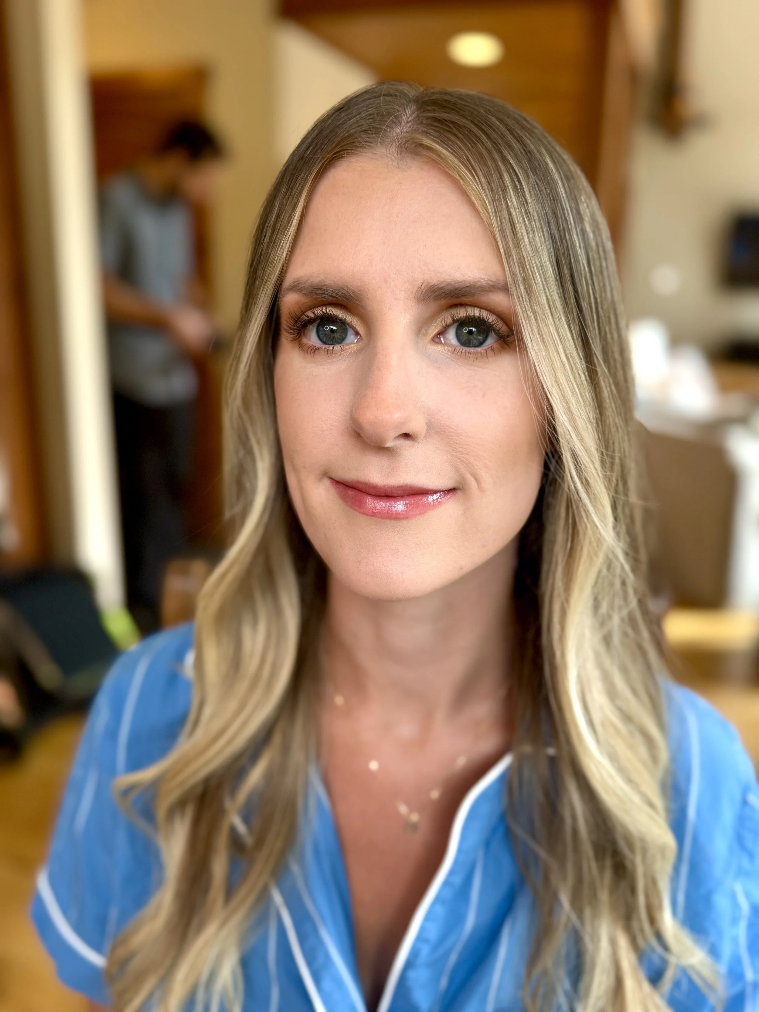 Close-up of a young woman with long blonde hair, wearing blue pajamas with white piping, and a delicate necklace, smiling softly, in a warmly lit room.