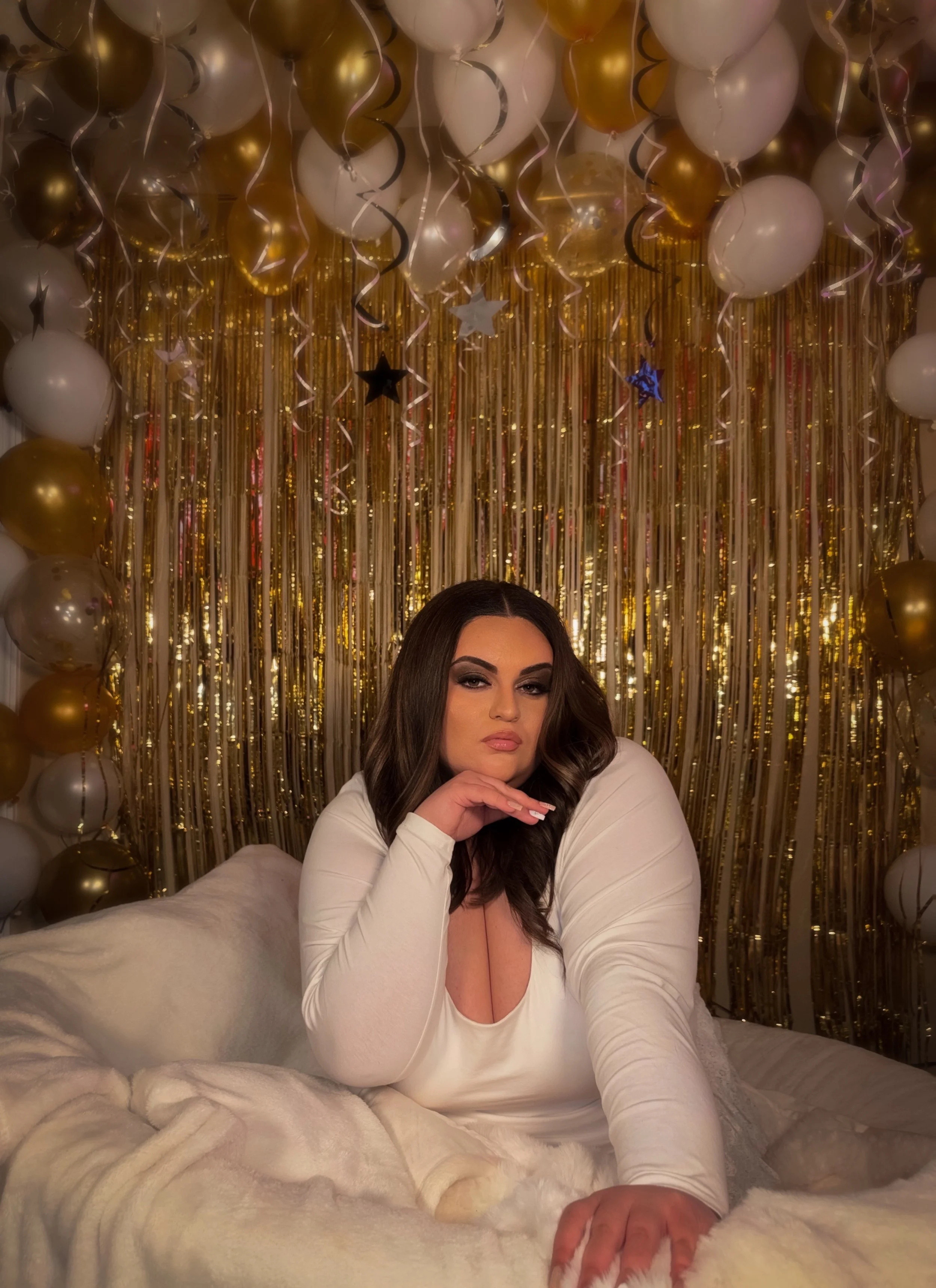 Woman with dark hair and makeup posing on a bed with a white fur blanket, in front of a gold and white balloon backdrop with hanging metallic streamers and star-shaped decorations, celebrating an event or party.