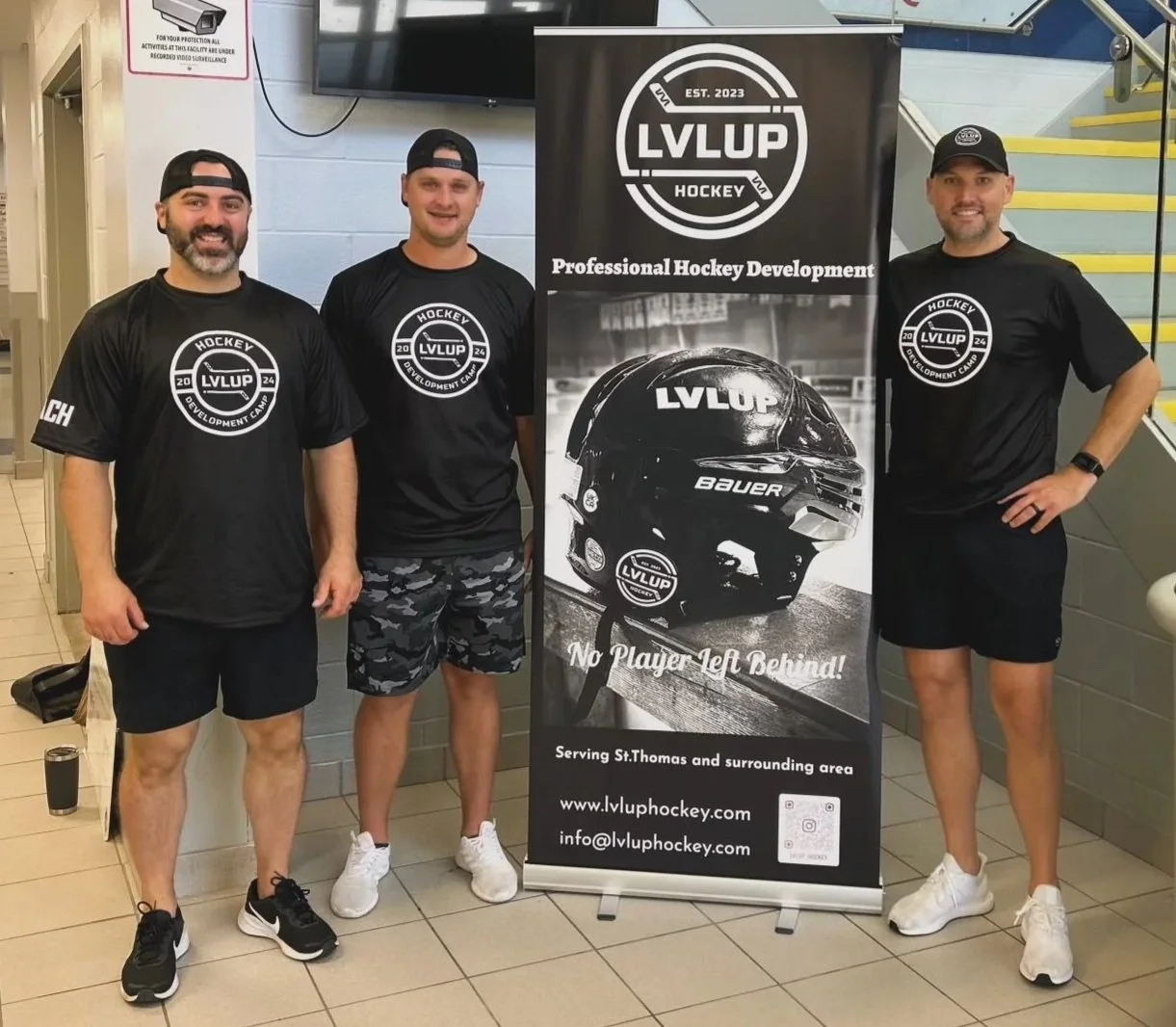 Three men standing indoors next to a vertical banner. All three are wearing black T-shirts with a circular logo for LVLUP Hockey Development Camp. The first man on the left has a beard and a backward cap, the second man in the middle is in camouflage shorts and a cap, and the third man on the right has a beard and a cap, with his hand on his hip. The banner features a hockey helmet with the LVLUP logo and text promoting professional hockey development, serving St. Thomas area, with website and contact email.