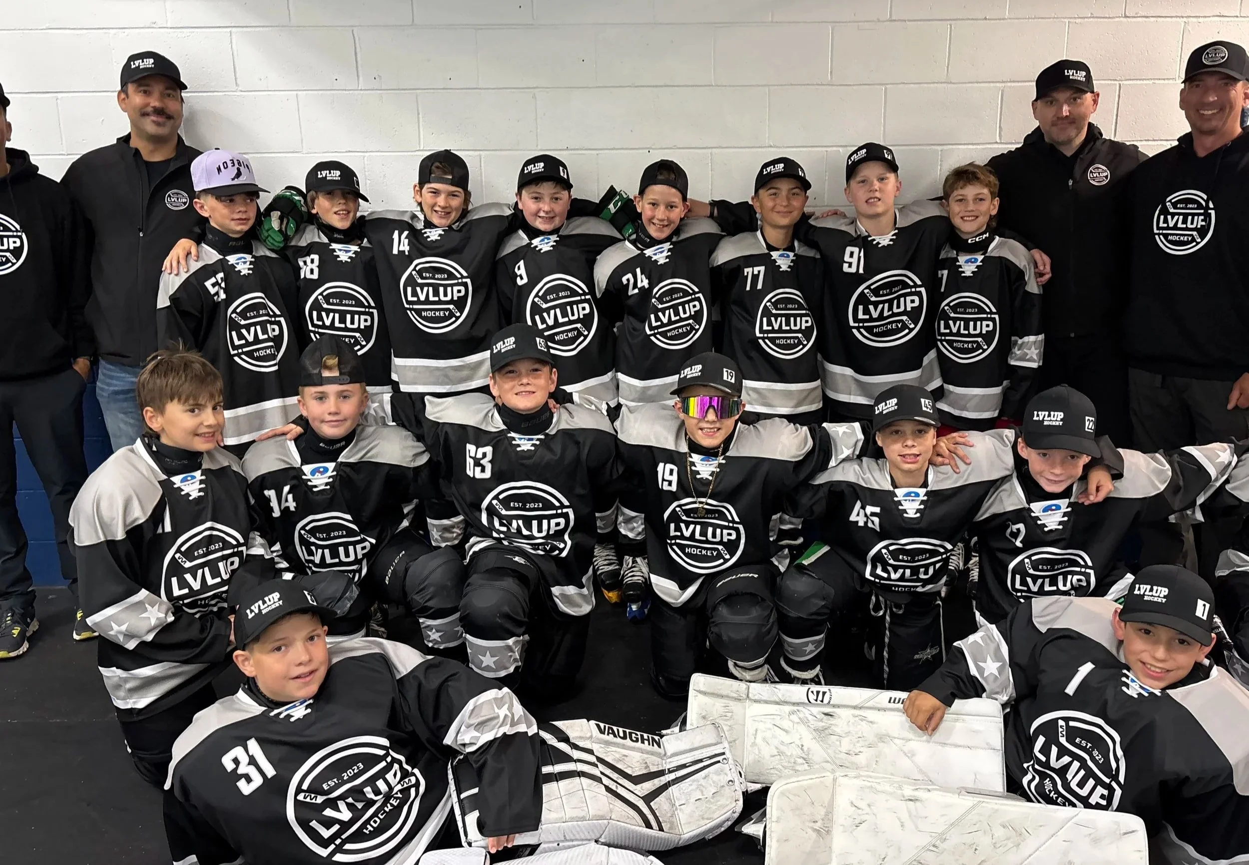 Group of young hockey players in black and grey jerseys with "LVLUP Hockey" logo, posing with coaches against a white wall.