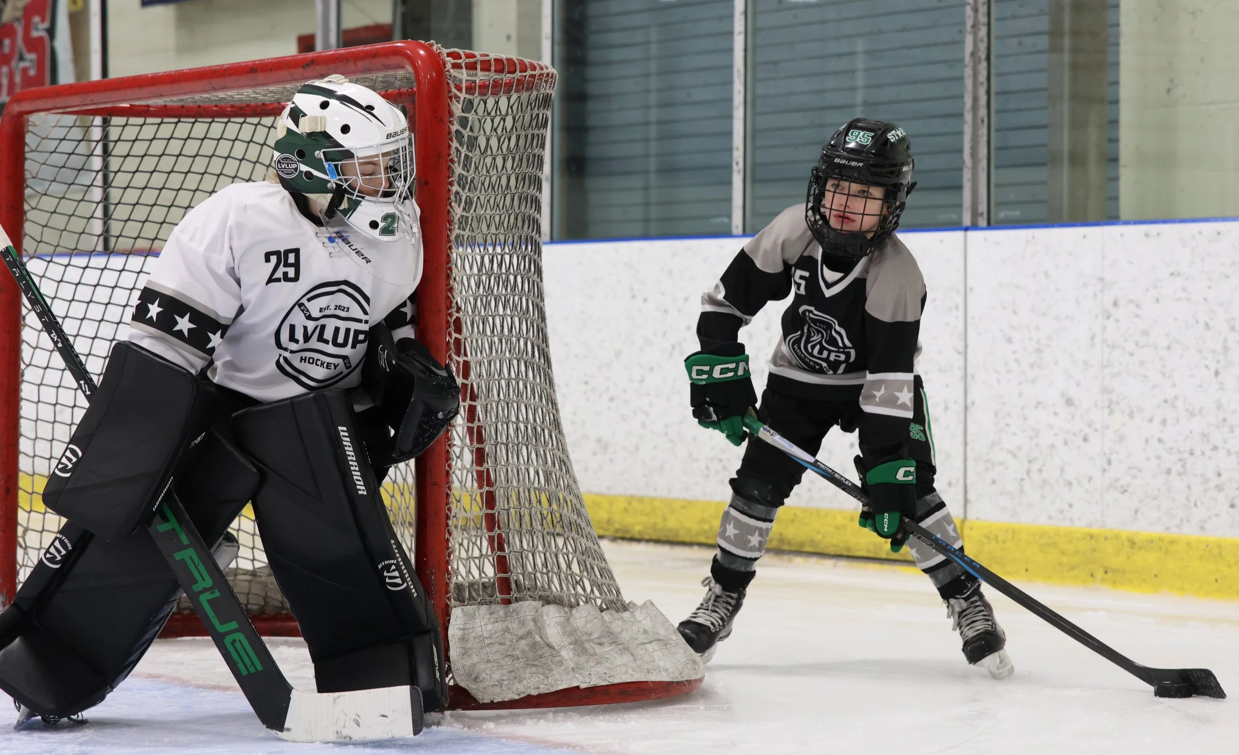A young hockey player in black and gray uniform with green gloves and helmet, standing in front of the goal, appears to be preparing to hit the puck. The goalie in white and black gear with a helmet marked with the number 29, is guarding the goal.