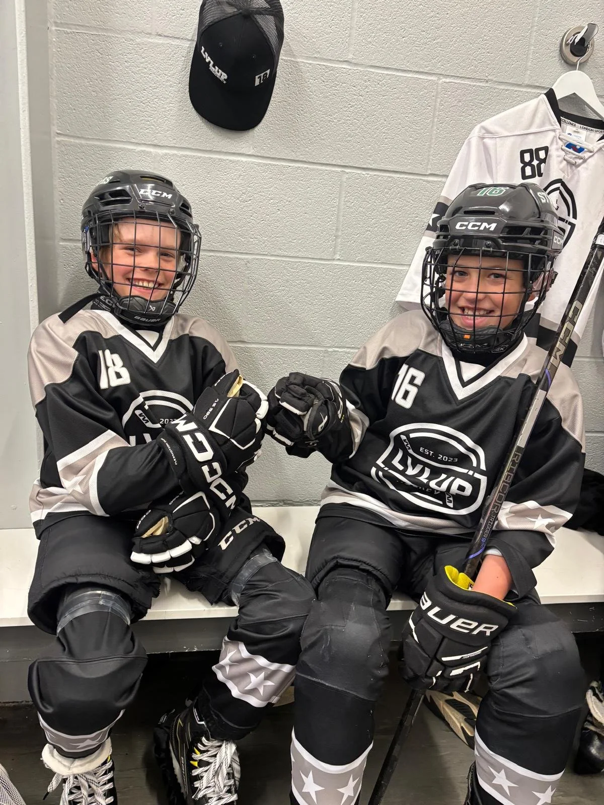 Two young boys in black and white hockey uniforms with helmets and gloves sitting on a bench, smiling, giving a fist bump, holding hockey sticks, in a locker room.
