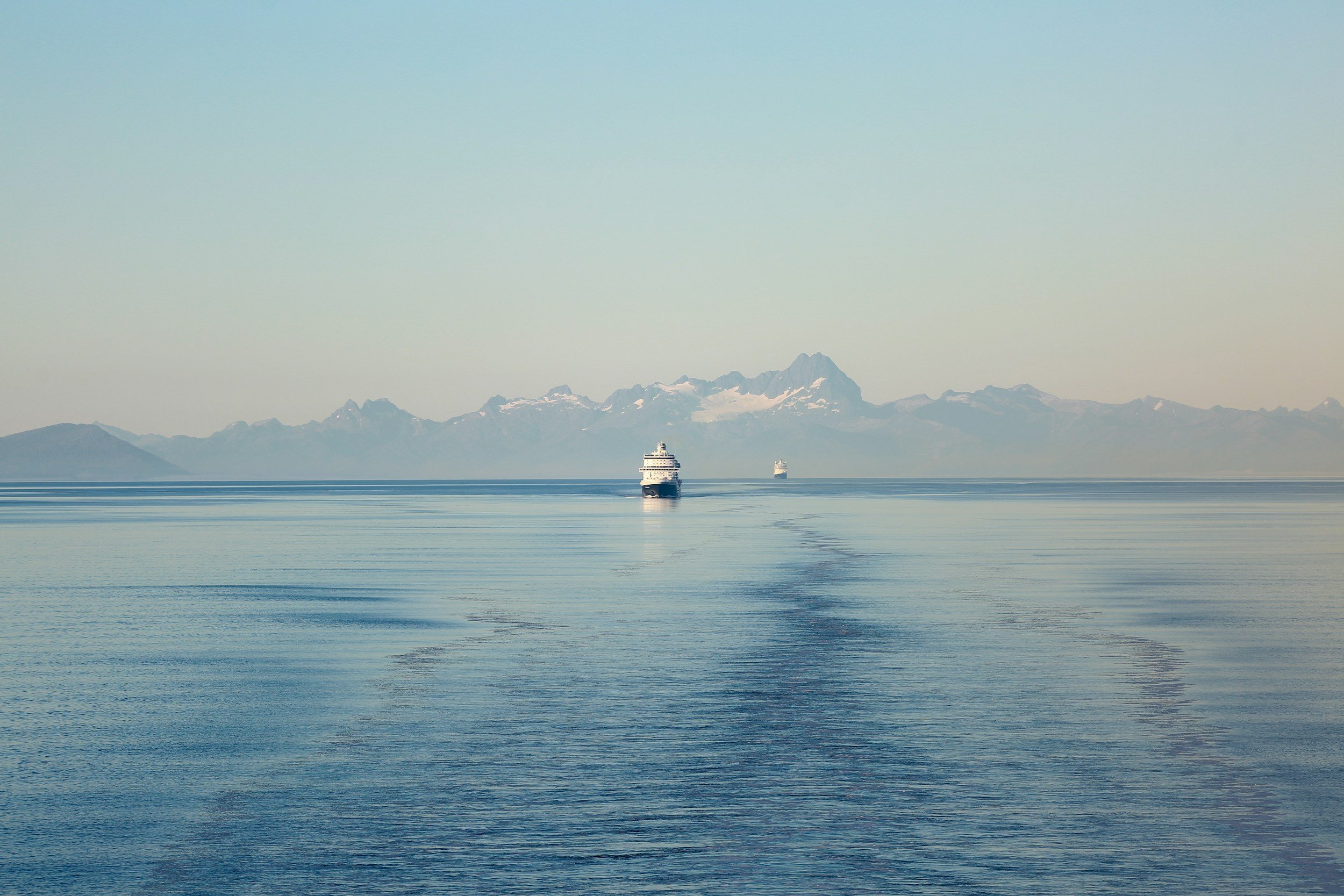 Two large ships sailing on calm ocean waters with snow-capped mountains in the background under a clear sky.