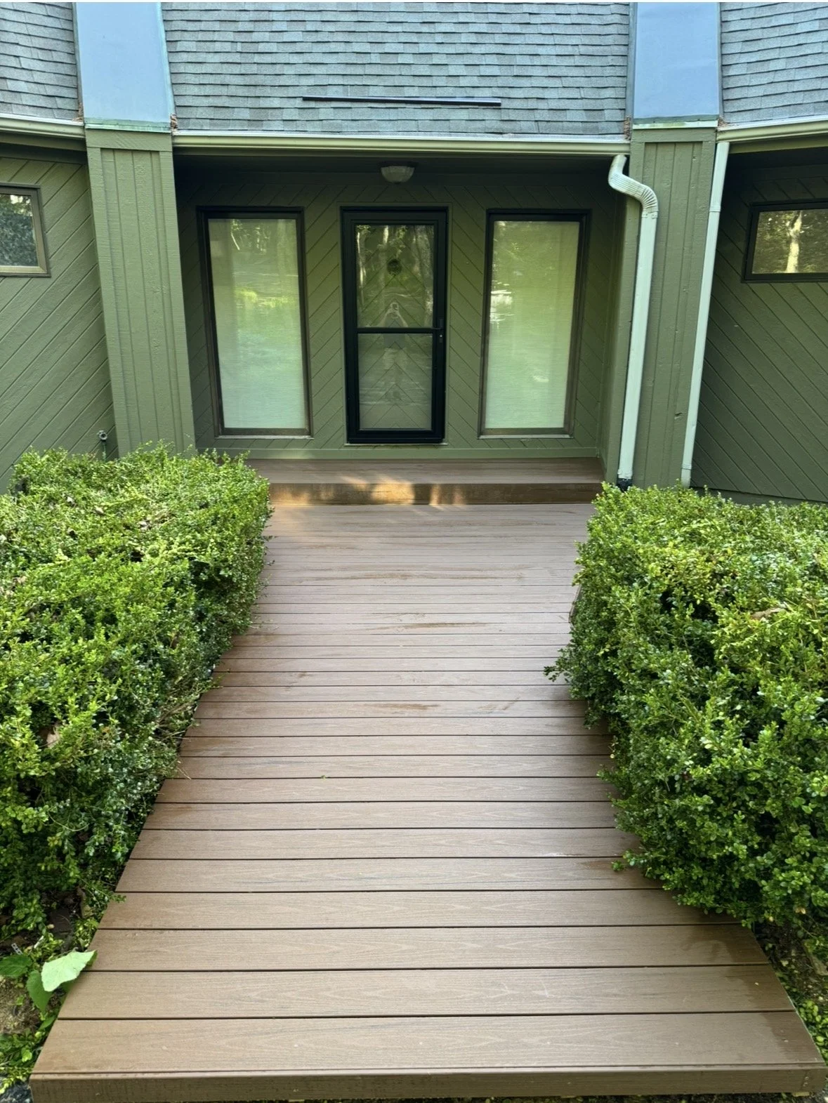 View of a wooden deck with bushes on each side, and a green house wall with a door and two tall windows.