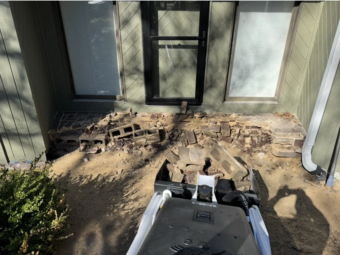 Construction site focusing on the front porch with scene of broken bricks and mortar, with a black door, windows, and a white downspout on the exterior of the house.
