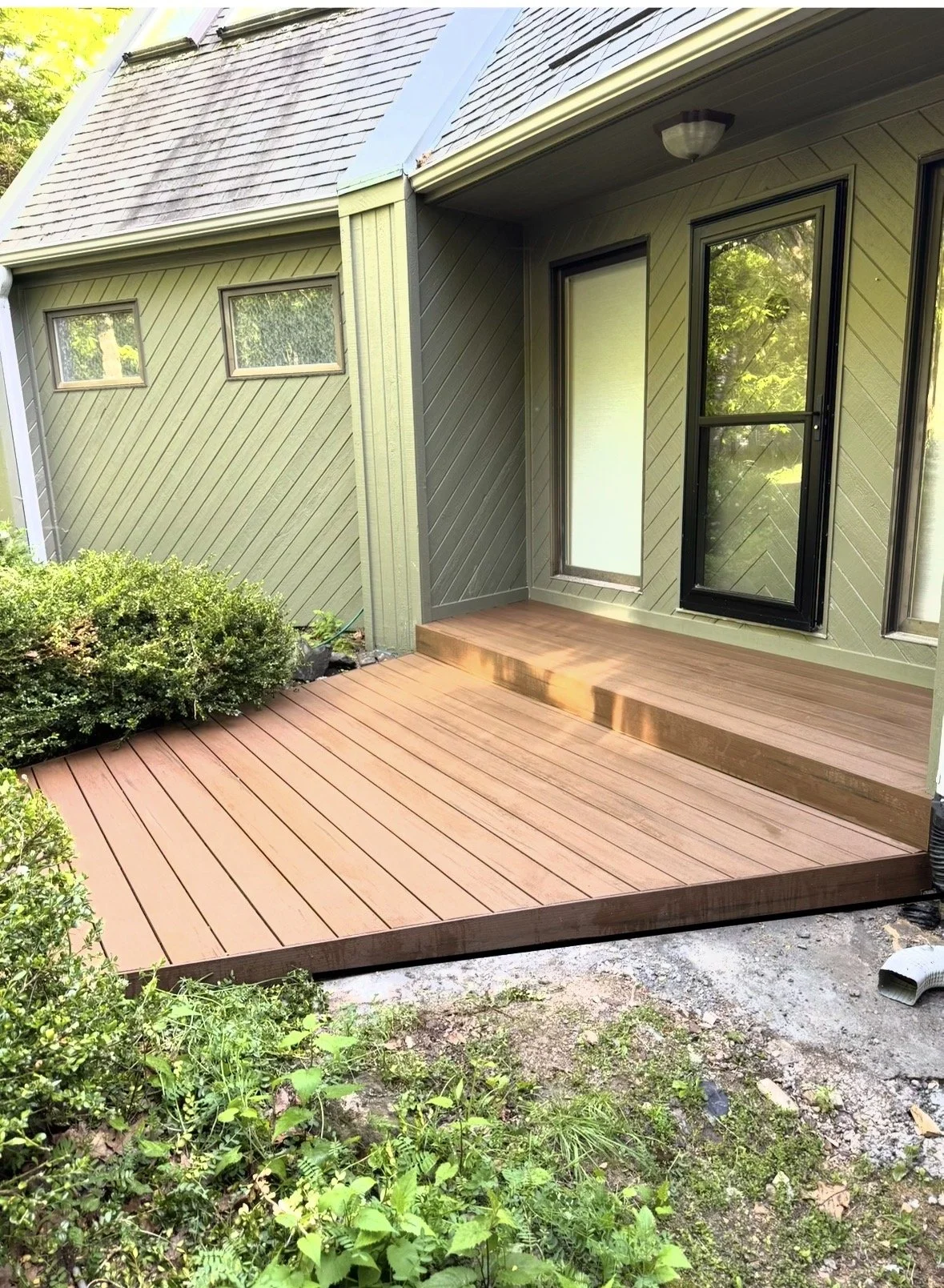 Newly built composite deck patio attached to a house with green siding, steps leading up to a glass door, surrounded by lush greenery.