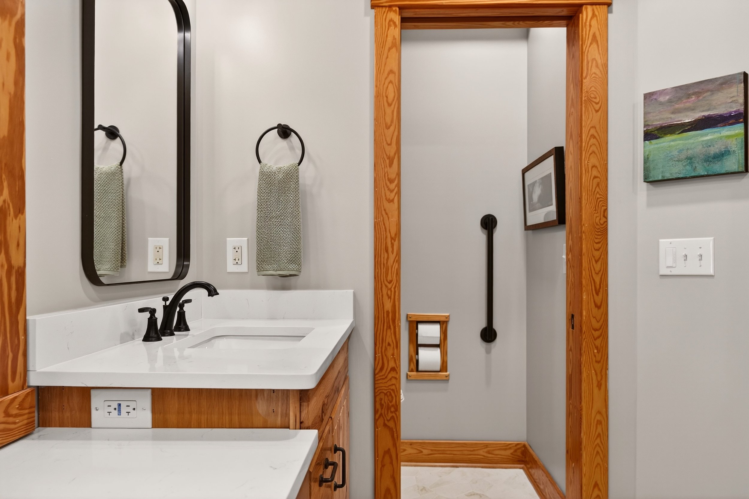 Bathroom vanity with a white countertop, black faucet, and mirror, framed in black, mounted on a white wall. There are two electrical outlets below the counter. A beige hand towel hangs on a black ring next to the mirror. A wooden door frame with a m