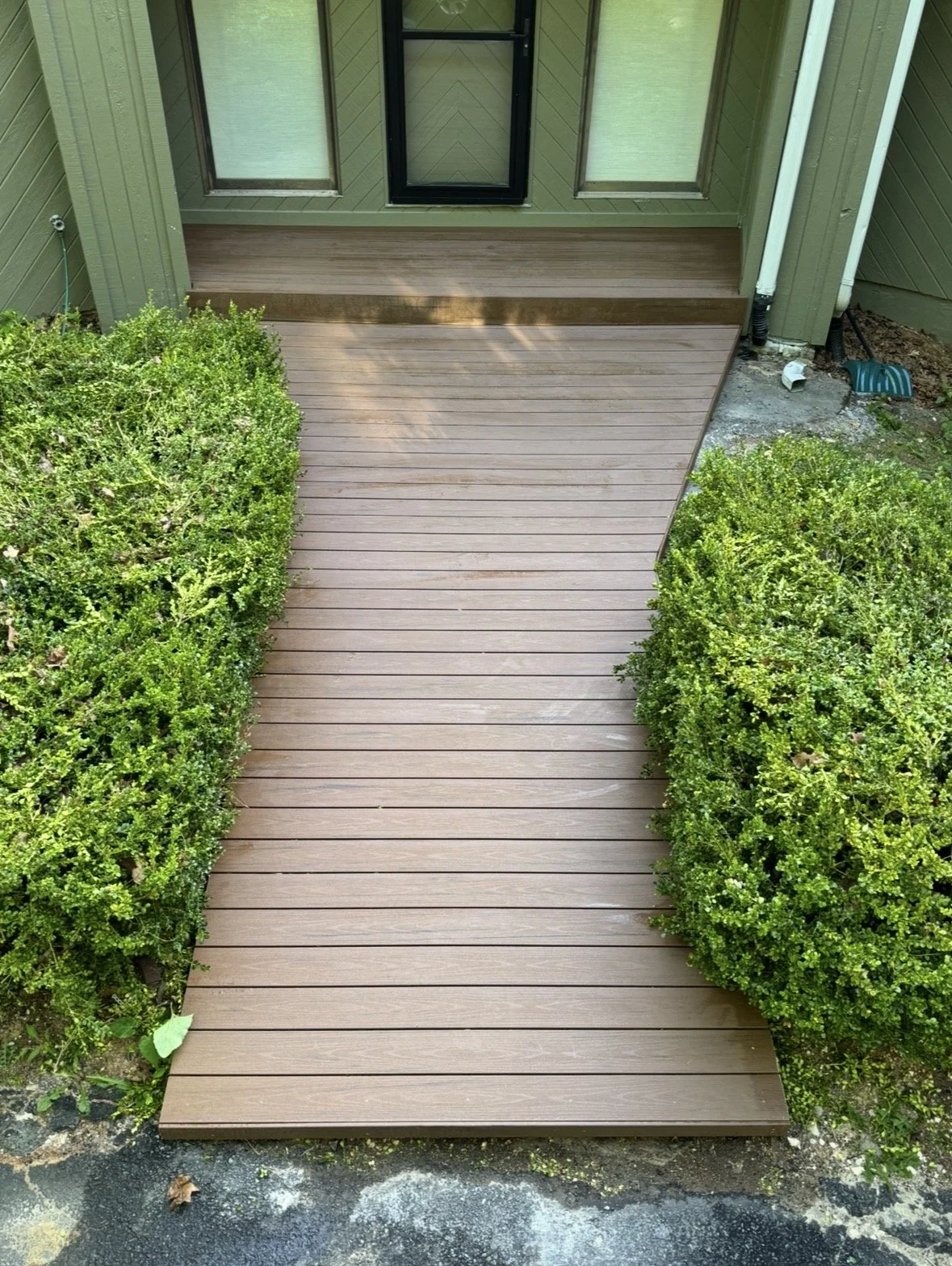 Newly installed brown wooden deck with green shrubs on either side, outside a house with green siding, windows, and a door.