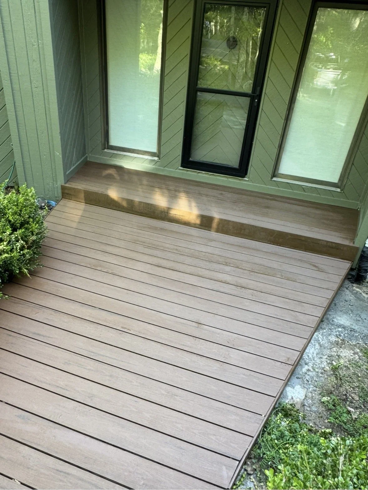 Newly installed composite deck with steps leading up to a screened porch with glass door and windows, surrounded by greenery.