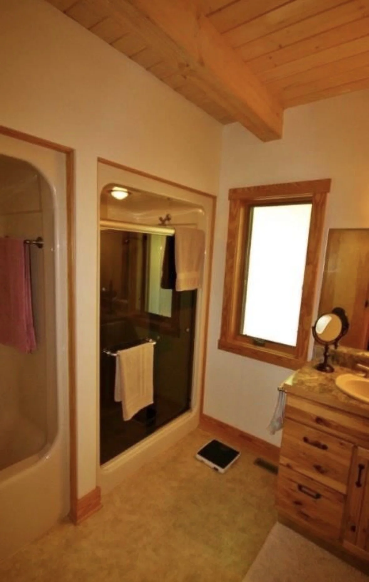 Interior view of a bathroom with wooden accents, a shower with a glass door, a window with wooden framing, a wooden dresser, a small round mirror, and a scale on the floor.