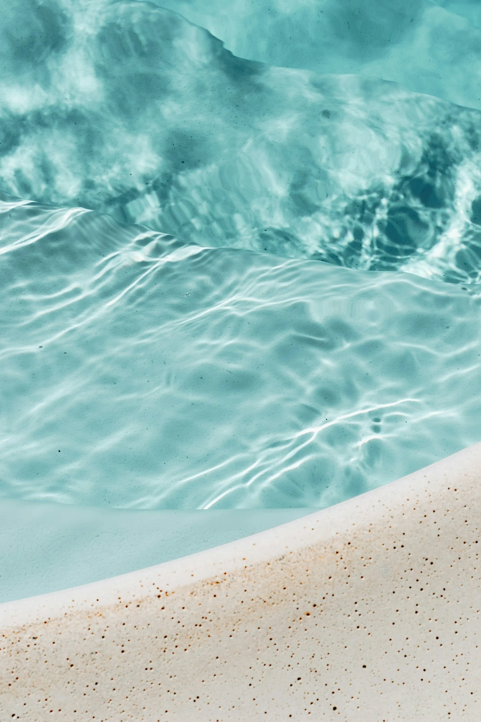 Close-up of clear, turquoise swimming pool water with sunlight reflections, sand at the pool's edge.