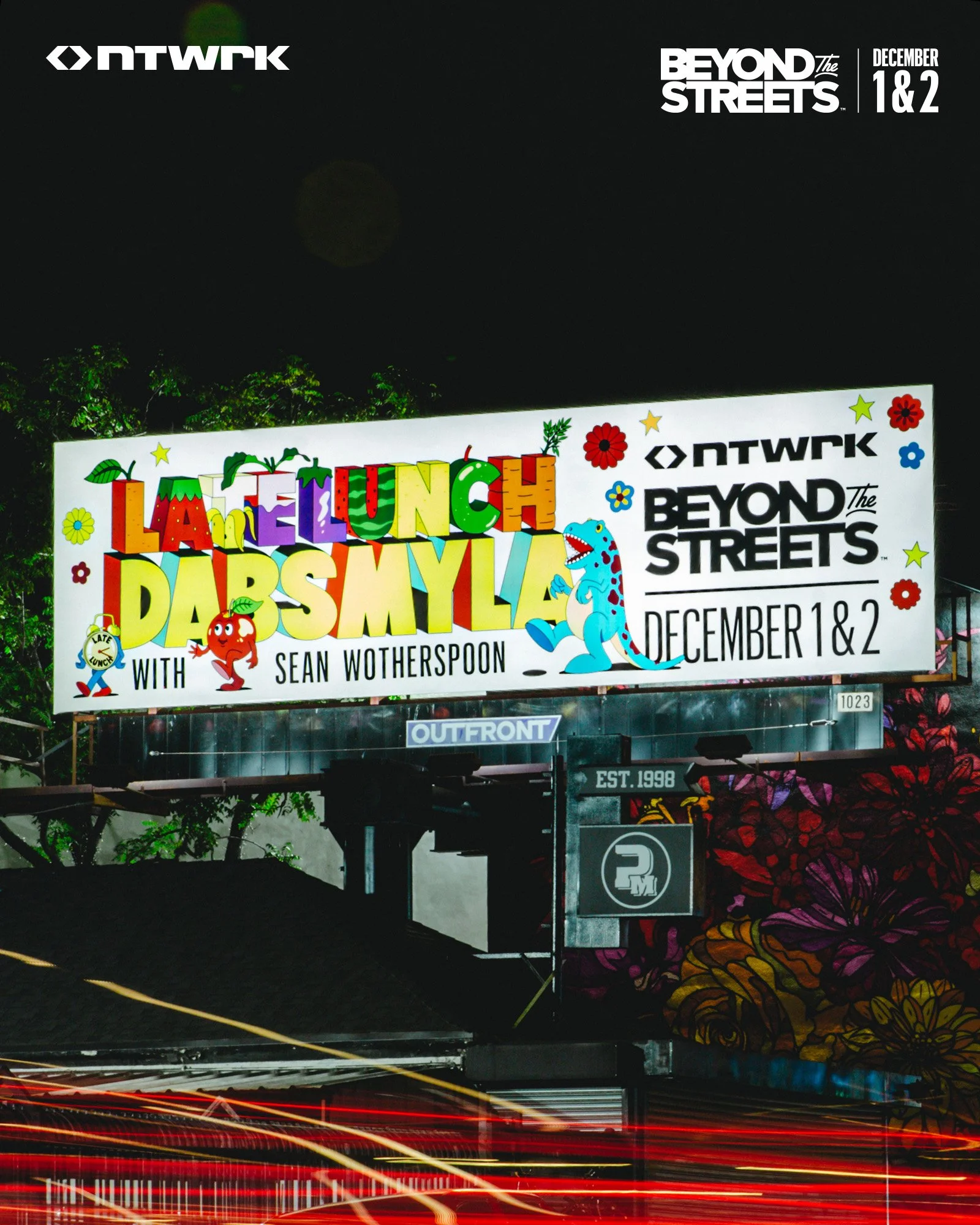 Bright billboard advertising an event called 'Late Lunch Dabs Myla with Sean Wotherspoon' at Beyond the Streets on December 1st and 2nd, 2023, featuring colorful cartoon illustrations of vegetables and dinosaurs over a night city backdrop.