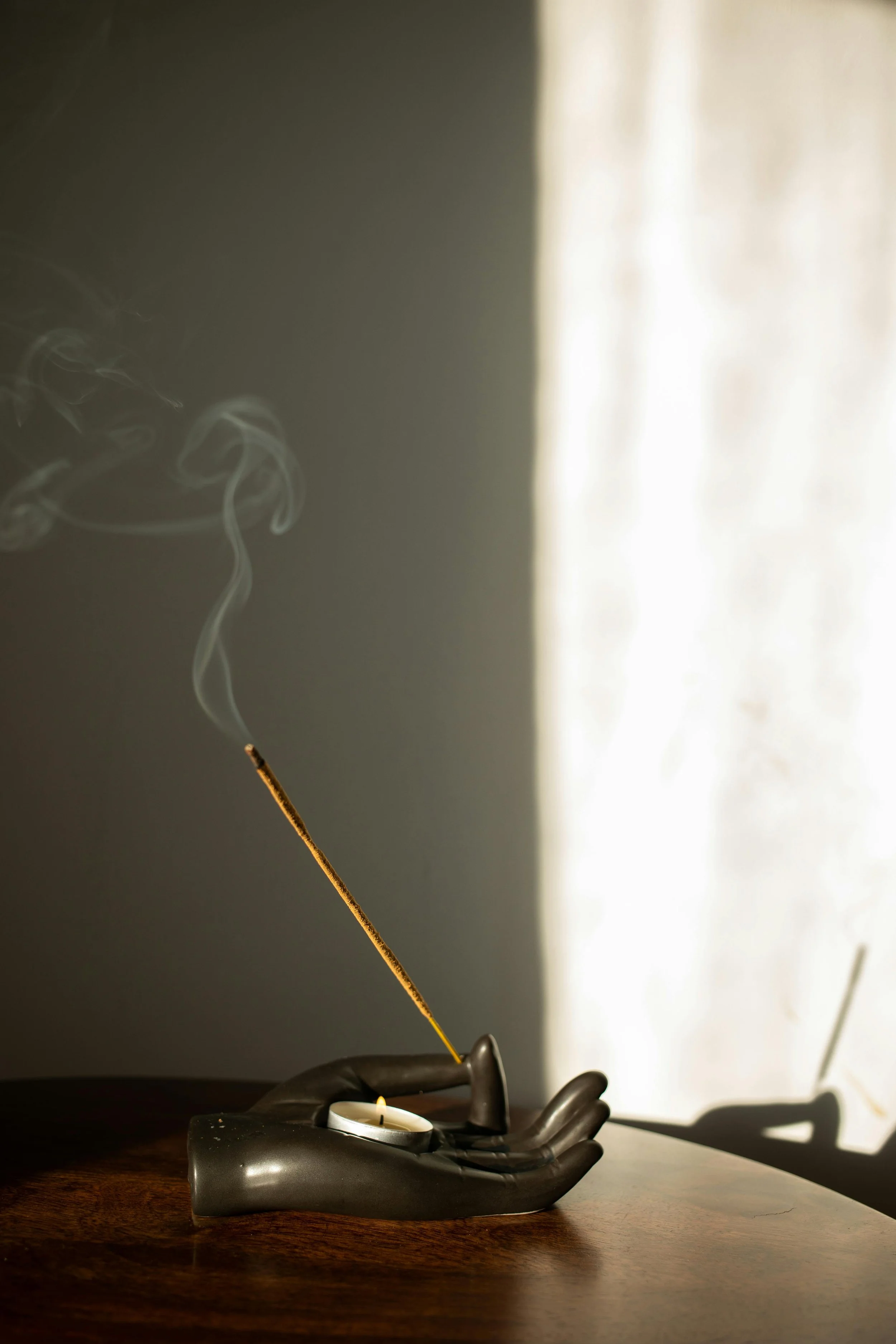 A moka pot in the shape of a human hand holding a tray with a tea light candle, a burning incense stick, and some fallen leaves on a wooden surface, with a shadow and light contrasting background.
