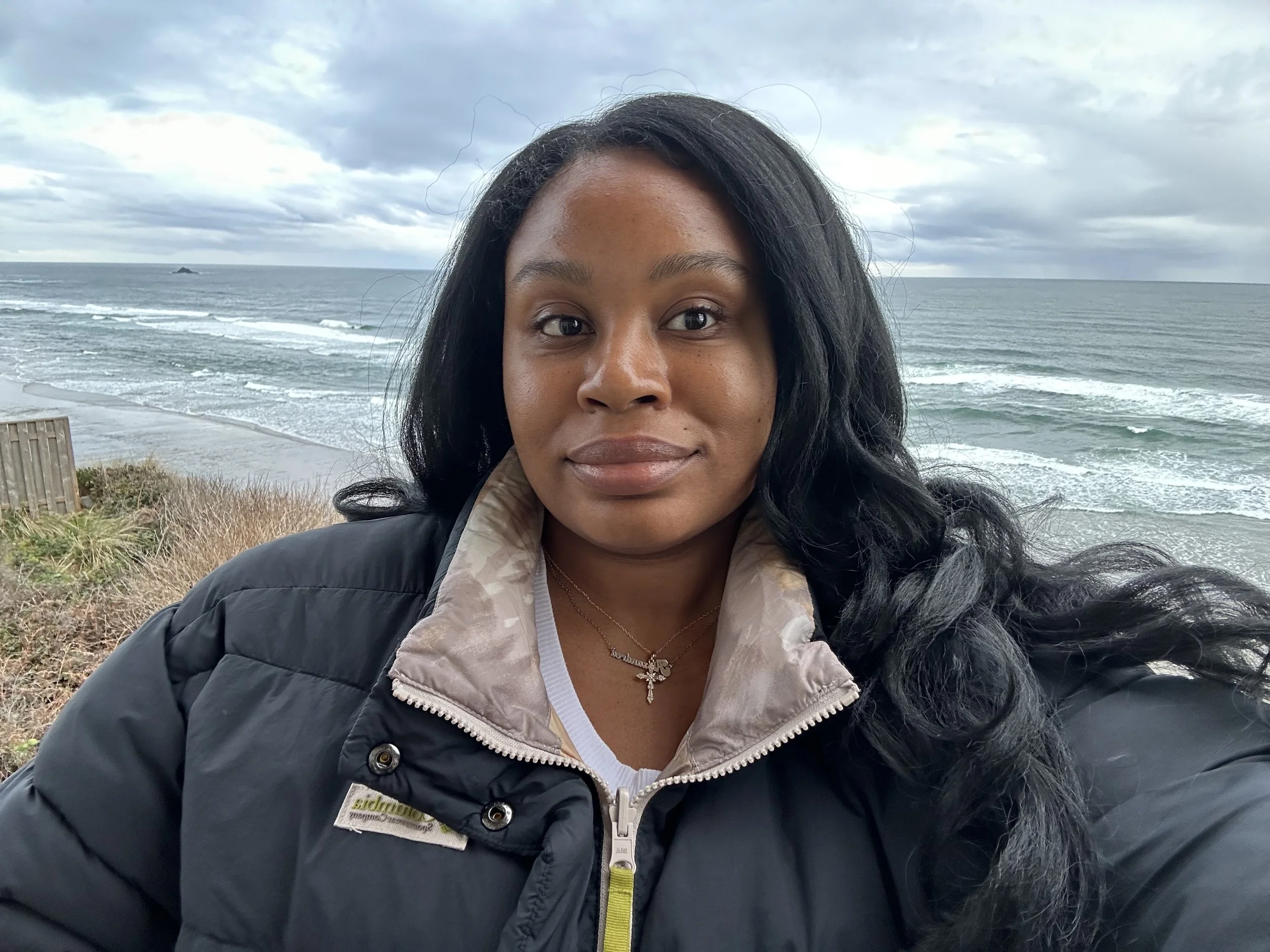 A woman with dark hair and wearing a black jacket with a beige collar, standing outdoors near the ocean on a cloudy day.