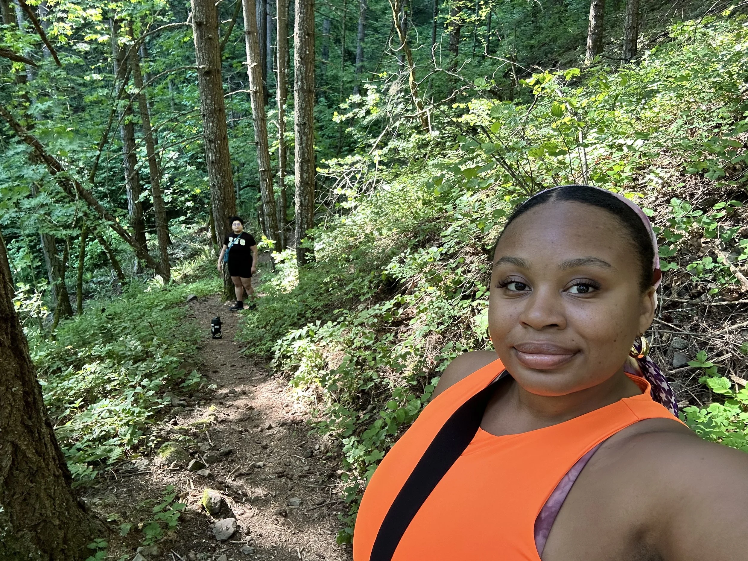 Close-up of a smiling woman with dark hair, wearing an orange top, taking a selfie on a forest trail with trees and green foliage in the background, and a person walking in the distance.