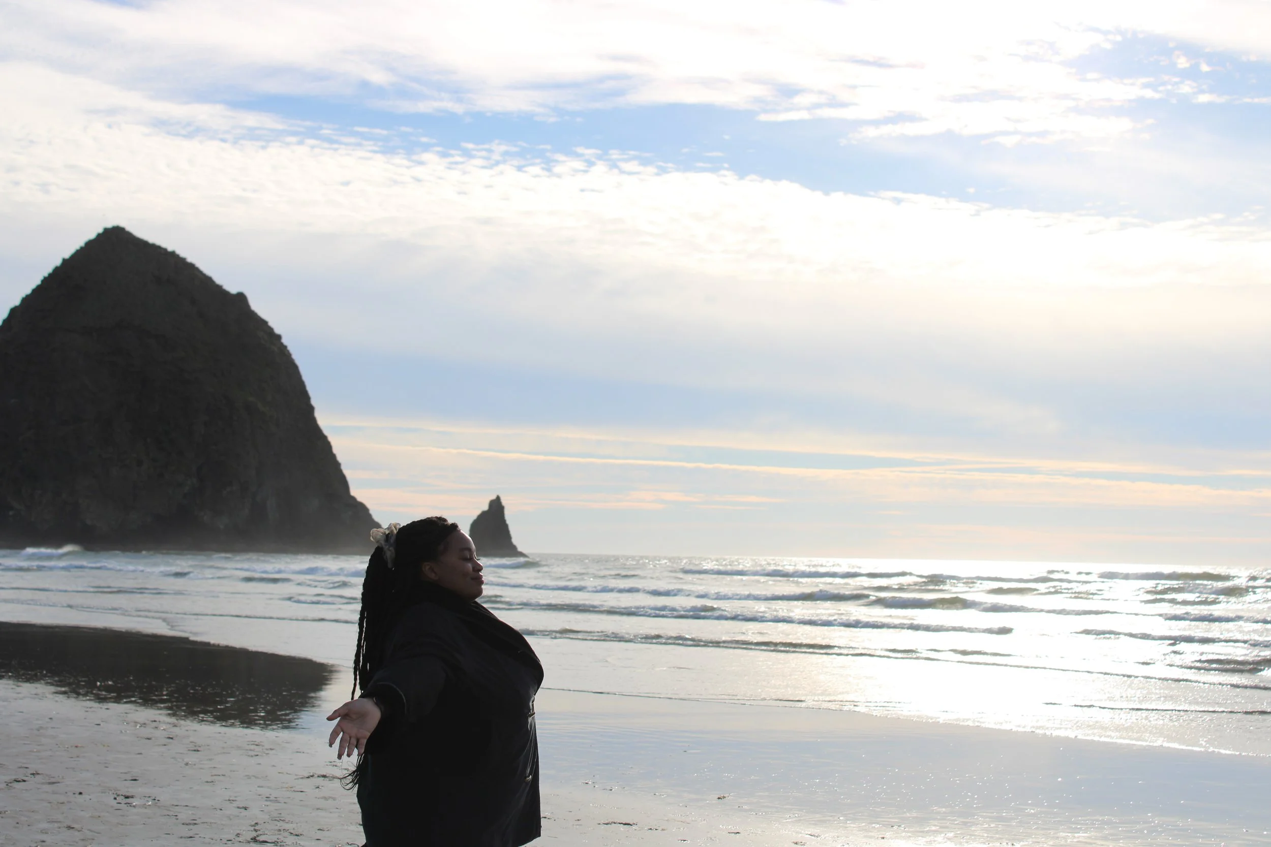 A woman with breads and a bow in her hair is standing on a beach near the ocean, with her arms outstretched and eyes closed, enjoying the scenery and the breeze. In the background, there is a large rocky island and clouds in the sky.