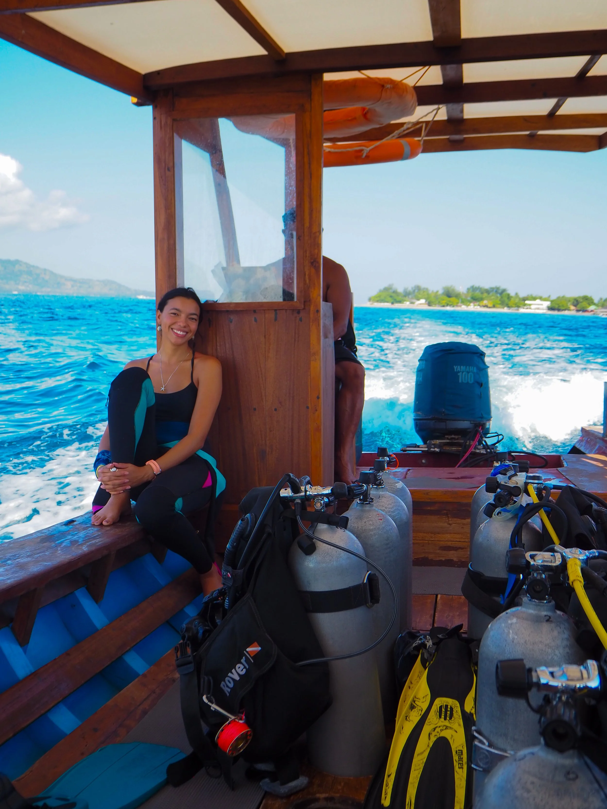 A smiling woman sitting on the edge of a boat with scuba tanks, equipment, and greenery in the background.