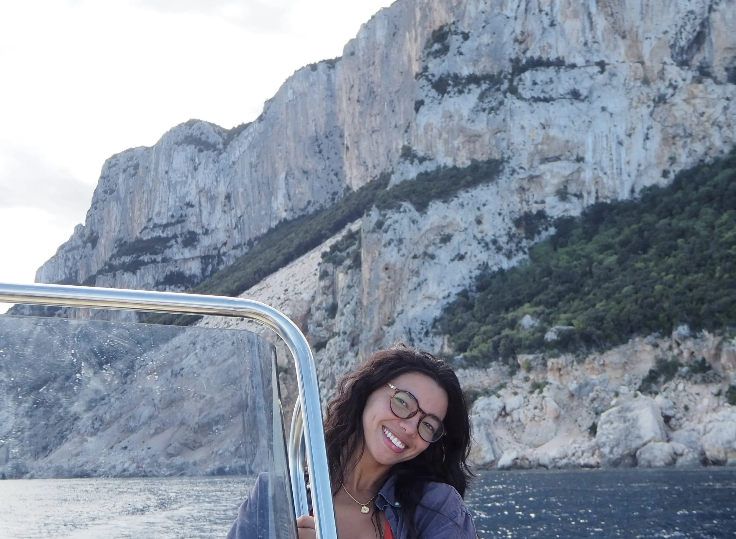 A woman with glasses smiling on a boat with a mountainous landscape in the background.