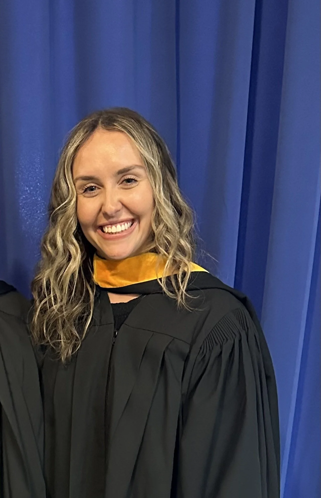 A woman in a graduation gown and hood, smiling, standing in front of blue curtains.