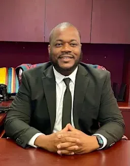 A man in a suit sitting at a desk with a bookshelf and maroon wall in the background.
