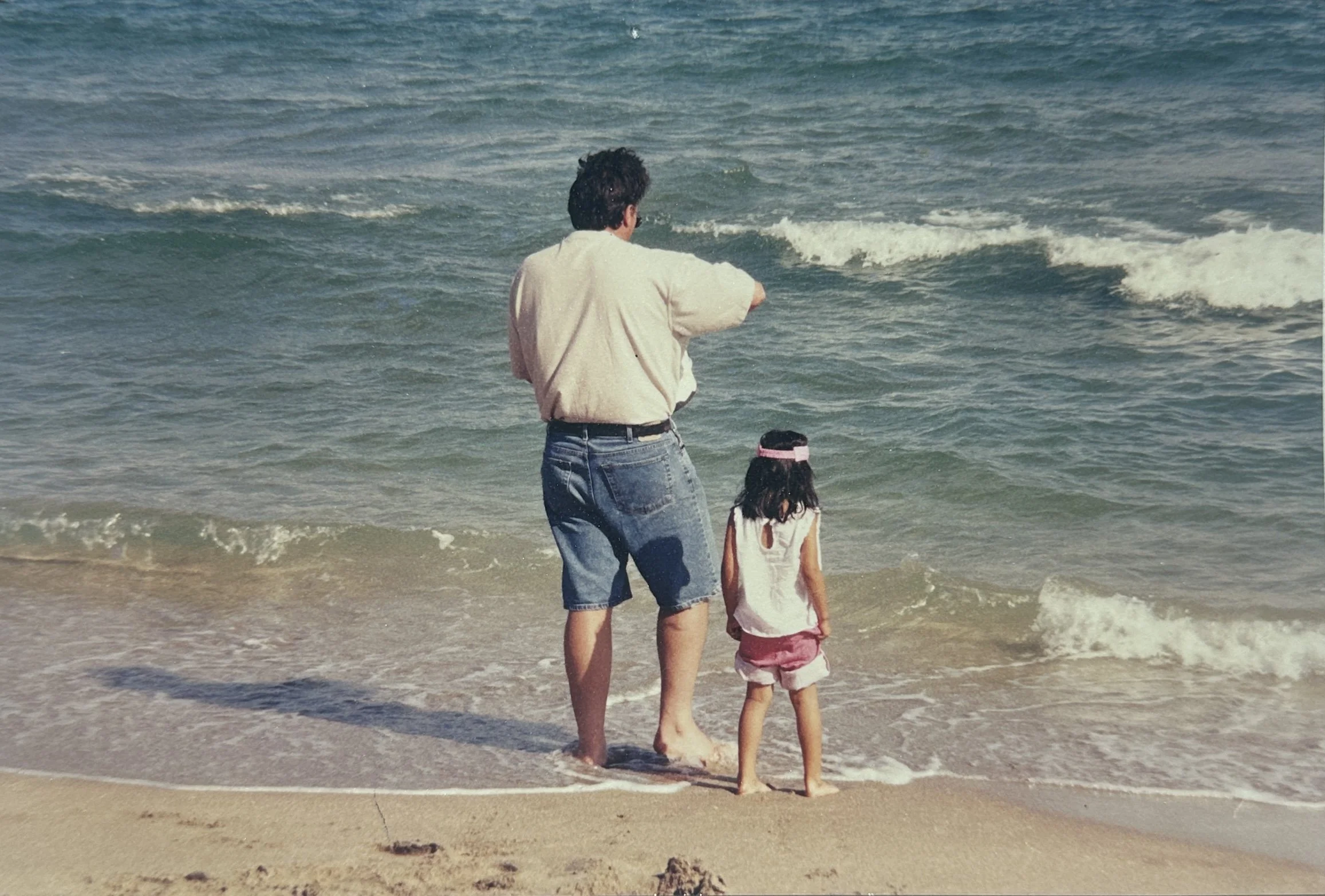 A man and a young girl walking on the beach near the water's edge, with small waves crashing nearby.