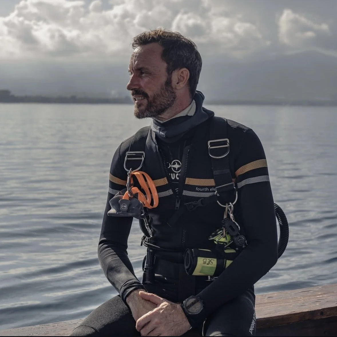 A man dressed in dive gear sitting by a body of water, with clouds in the sky behind him.