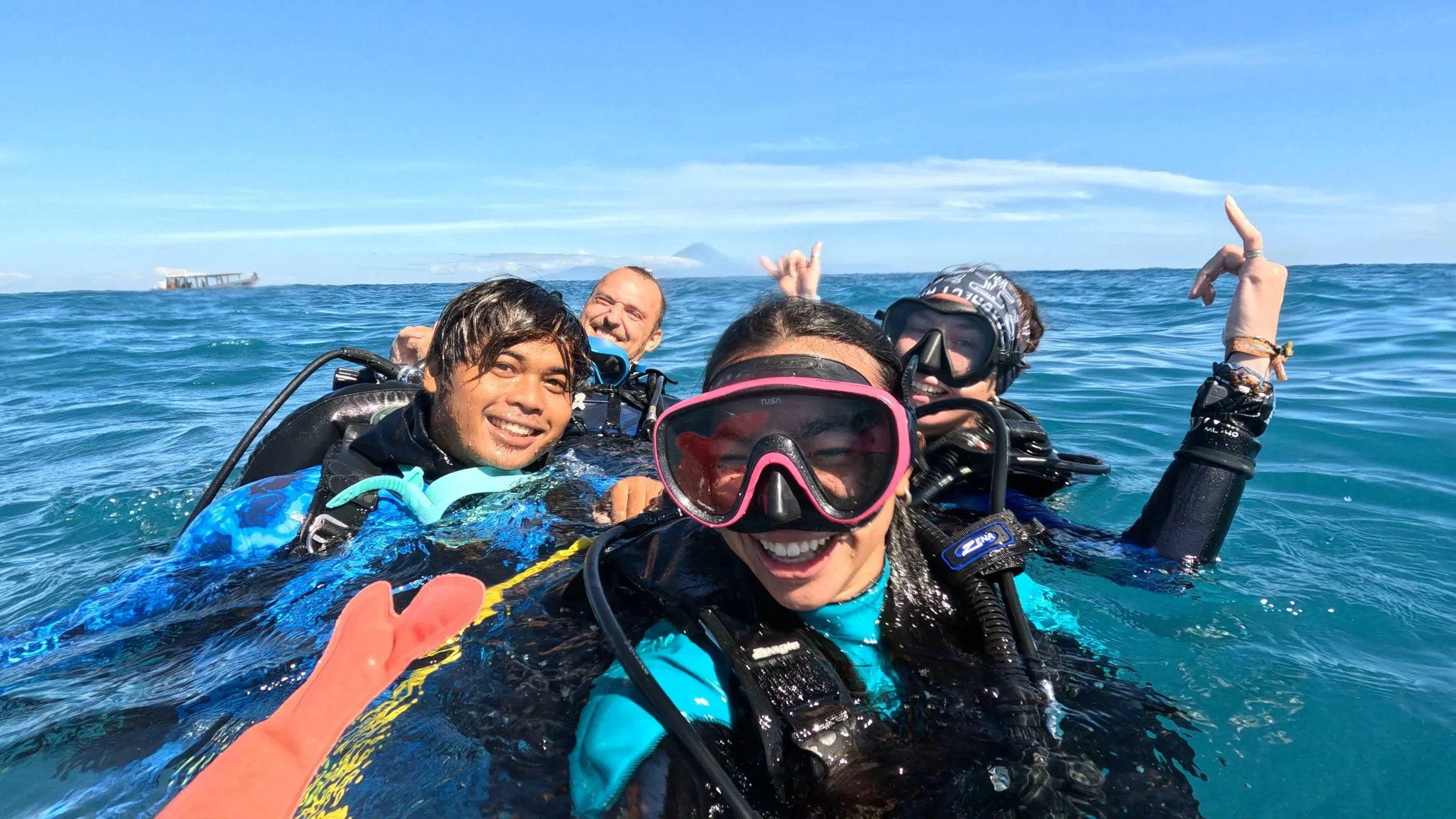 Four people in scuba gear smiling in the ocean, taking a selfie, with a ship in the background under a blue sky.