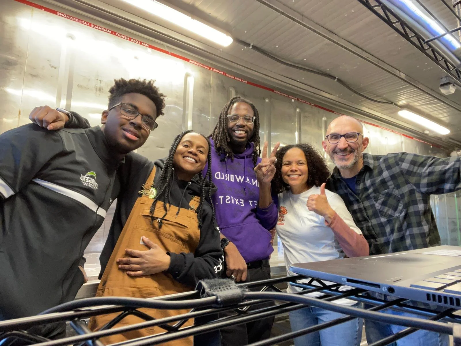 Group of five diverse people smiling and posing for a photo indoors, with a laptop and cables in the foreground, and a metallic wall background.