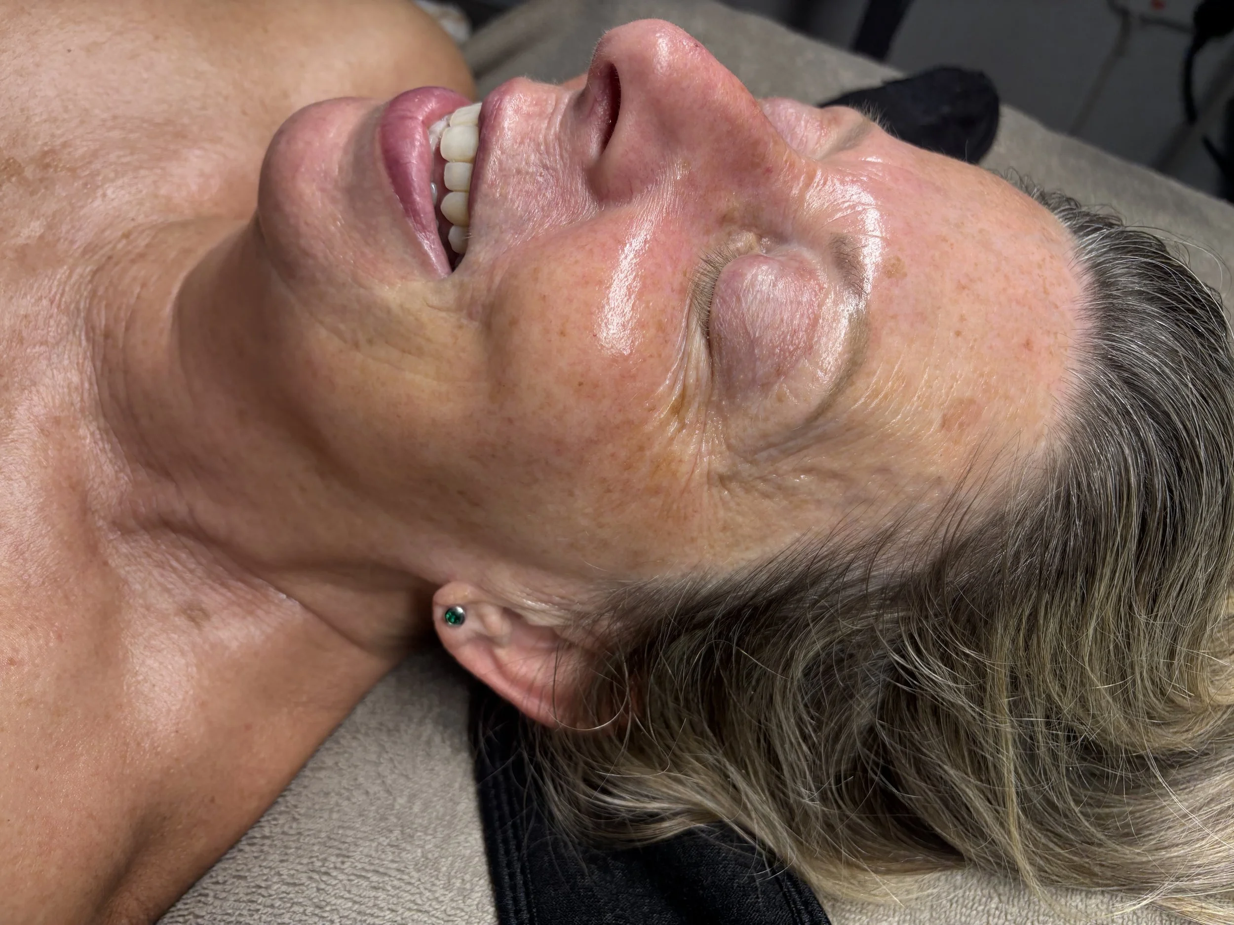 Close-up of a person's face with tanned skin, freckles, gray hair, and a small sparkly earring, lying down with eyes closed and smiling.