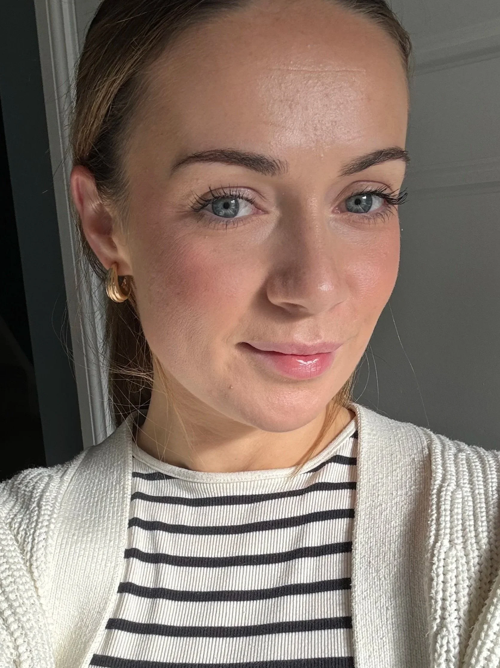 Close-up of a young woman with blonde hair, blue eyes, wearing gold hoop earrings, a white cardigan, and a black and white striped shirt, standing indoors near a door.