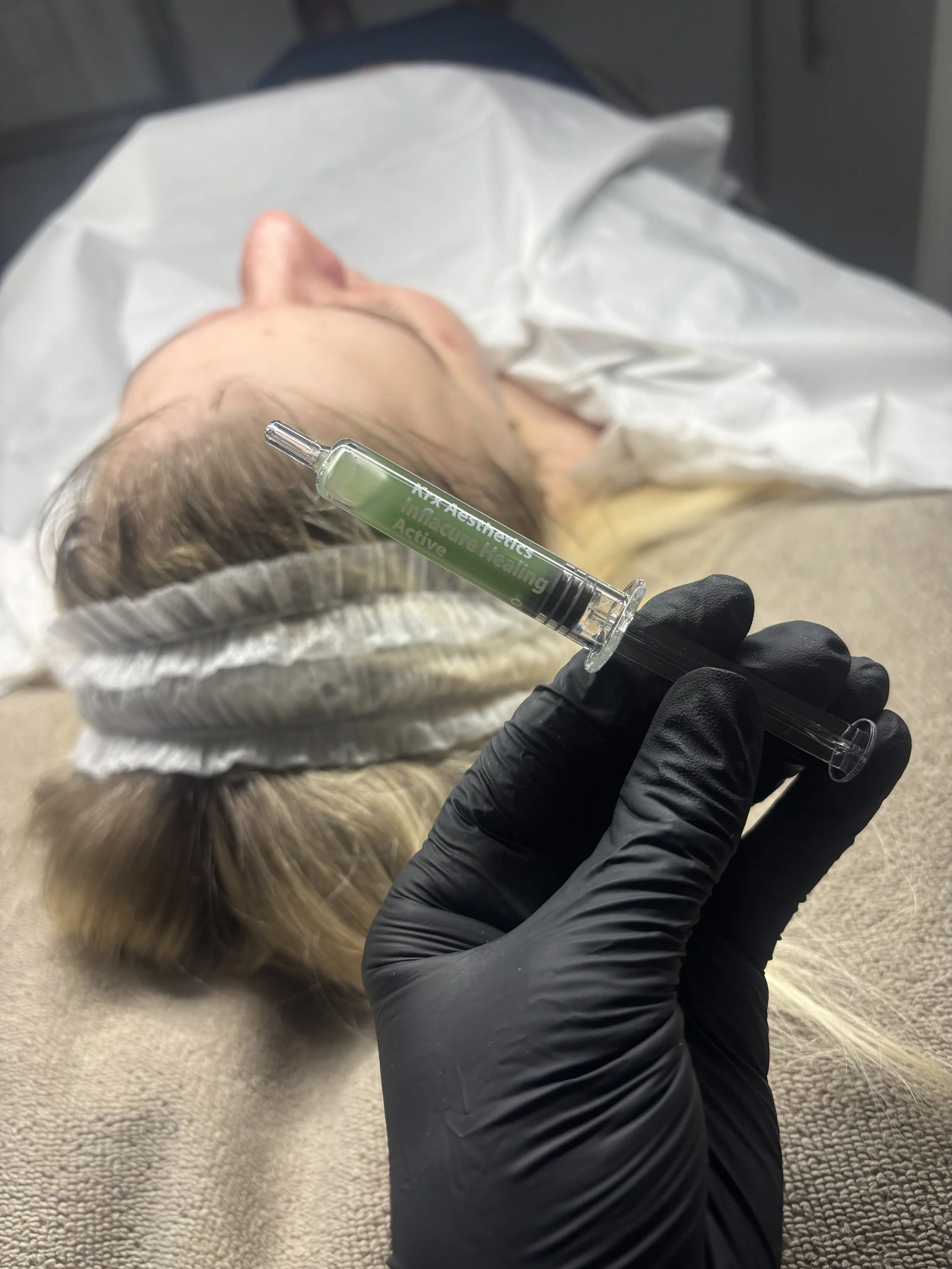 A person lying on their back on a medical bed, with a gloved hand holding a syringe filled with a clear substance labeled 'Immacure Healing Active' near their head.