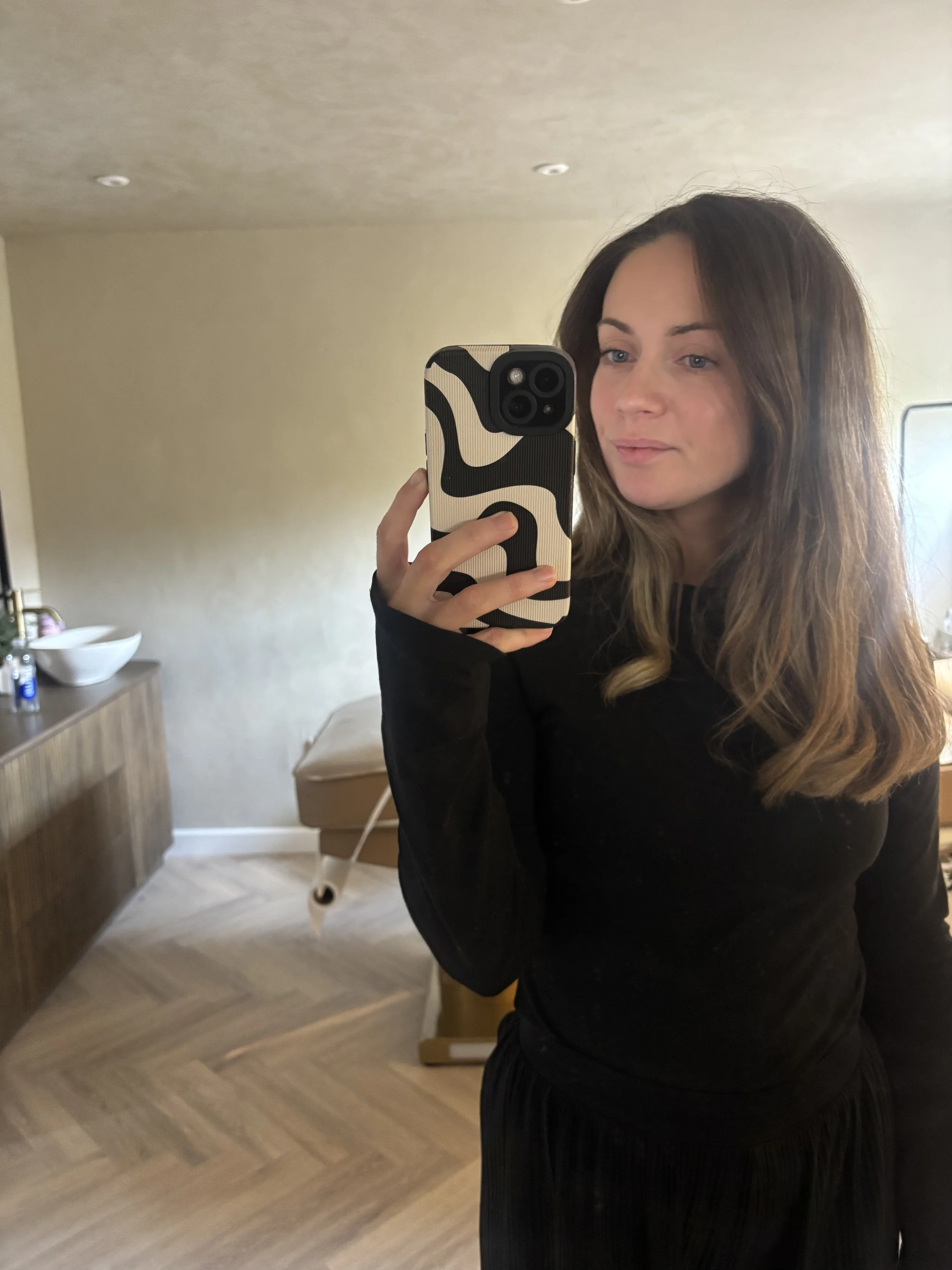A woman takes a mirror selfie in a room with wooden floors and beige walls, holding a phone with a black and white abstract patterned case. She has long, wavy brown hair and is wearing a black top.