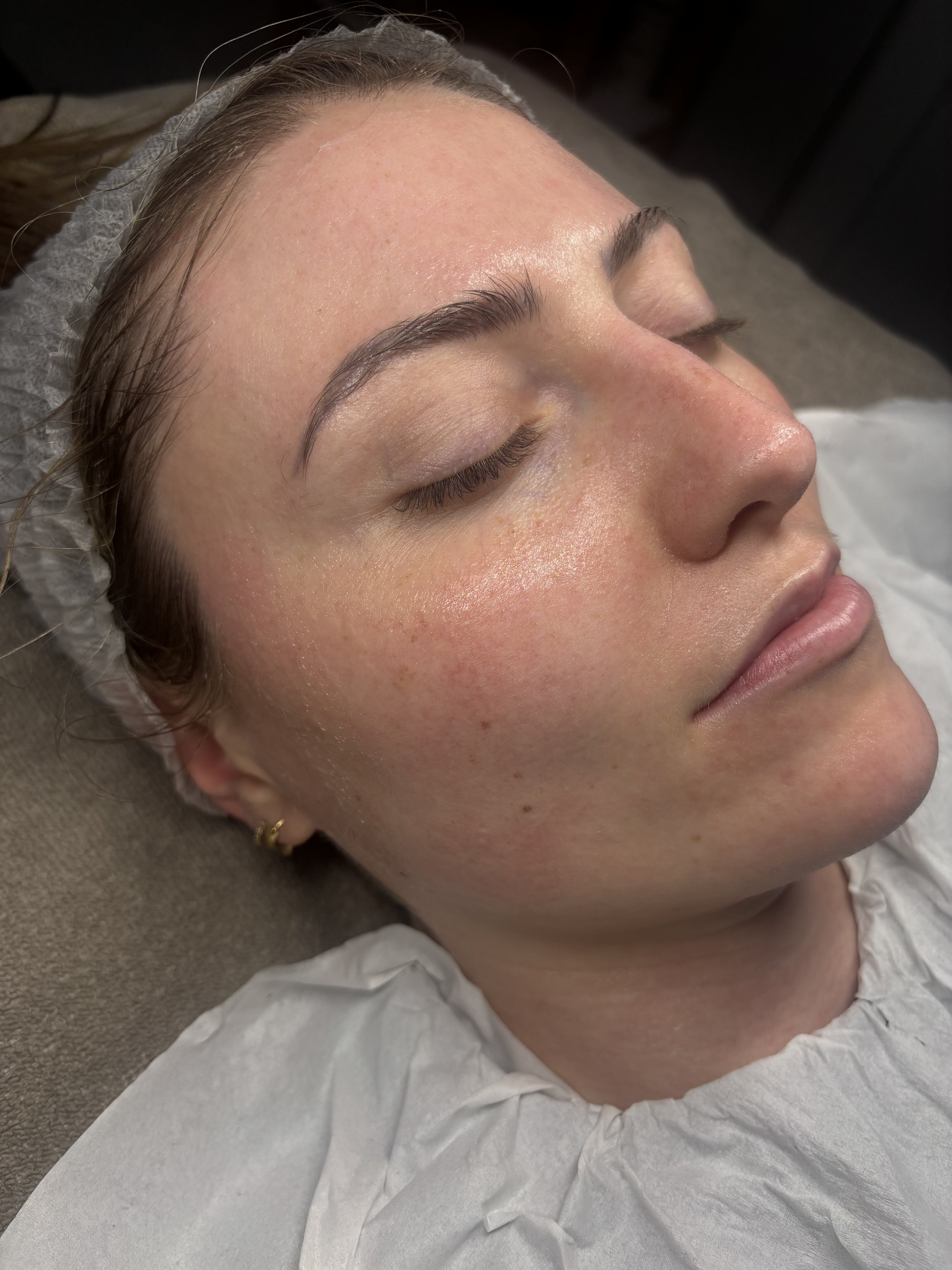 A woman is lying down with her eyes closed, appears to be relaxing or receiving a facial treatment, with her head on a cushioned surface, covered with a protective sheet.