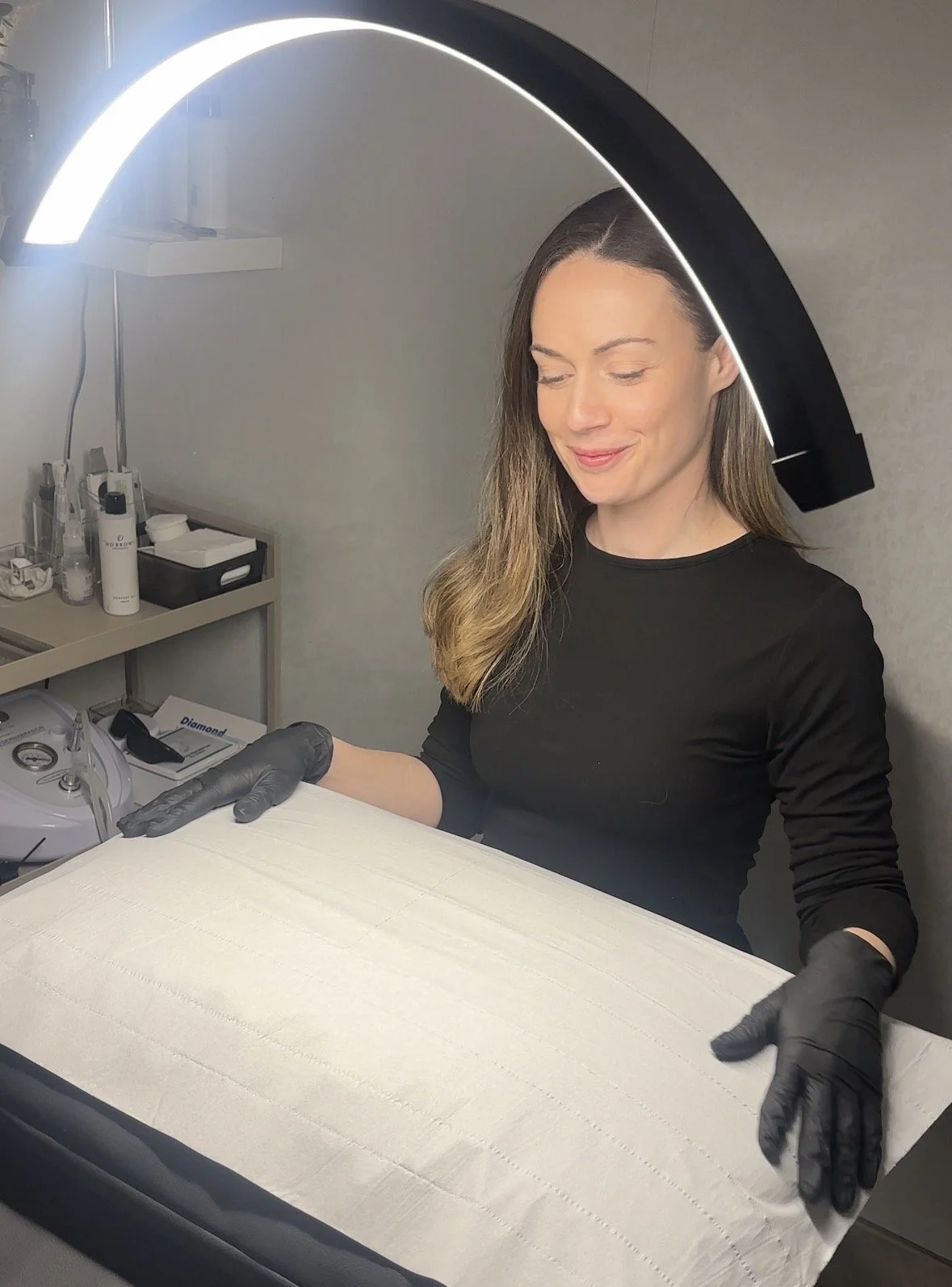 A woman wearing black gloves handling a white fabric surface in a medical or clinical setting, with various equipment and bottles on a shelf in the background.