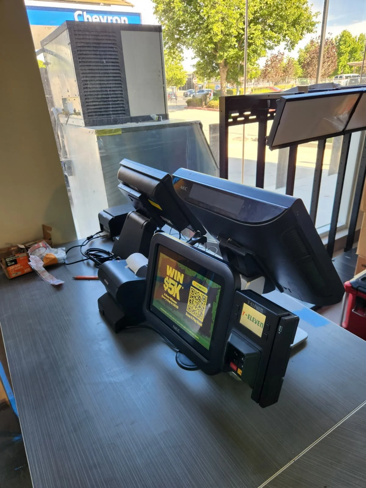 Point-of-sale register with a digital screen displaying a promotional offer, a cash drawer, a barcode scanner, and other cash register equipment on a retail counter near a window.