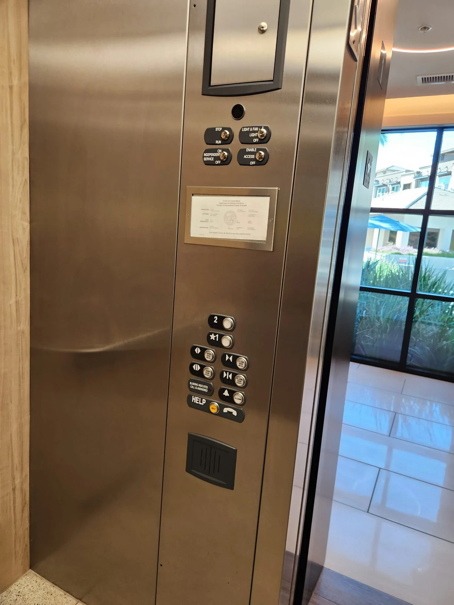 Interior view of an elevator control panel with various buttons, a screen, and a window showing a building outside.