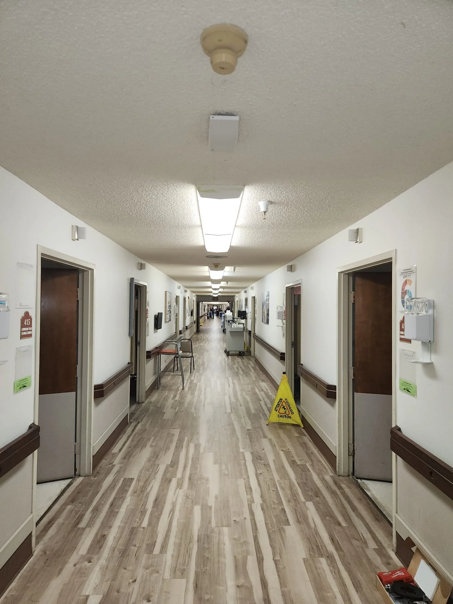 Hospital corridor with open patient room doors, chairs, medical equipment, and a "Caution Wet Floor" sign on the floor.