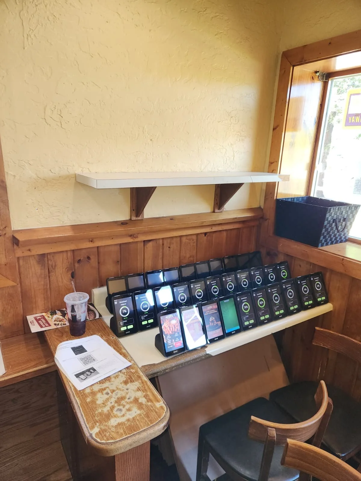 Multiple mobile devices placed on a table, displaying progress screens, inside a pizza restaurant. There is a drink cup and some papers on the table, with wooden walls and a window in the background.