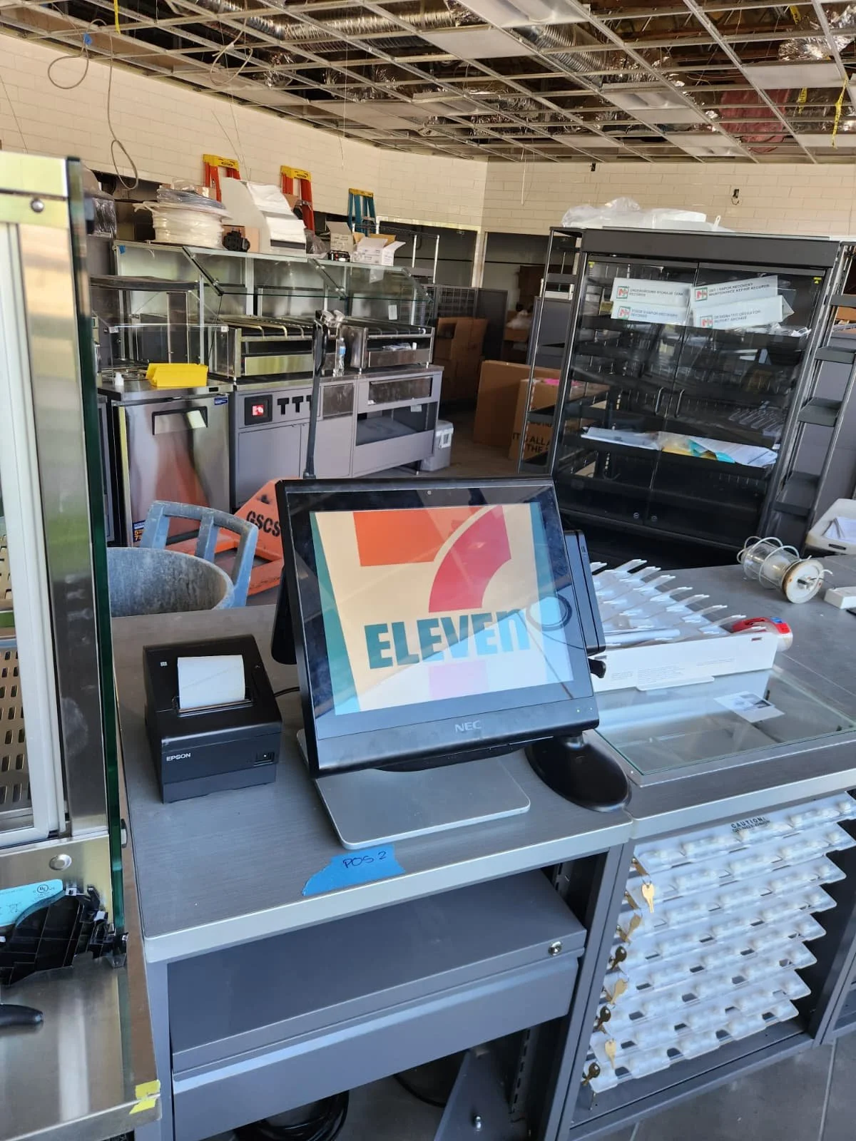 Cash register, receipt printer, and a monitor displaying the logo for Eleven, in a commercial kitchen or cafeteria setting with unfinished ceiling and equipment in the background.