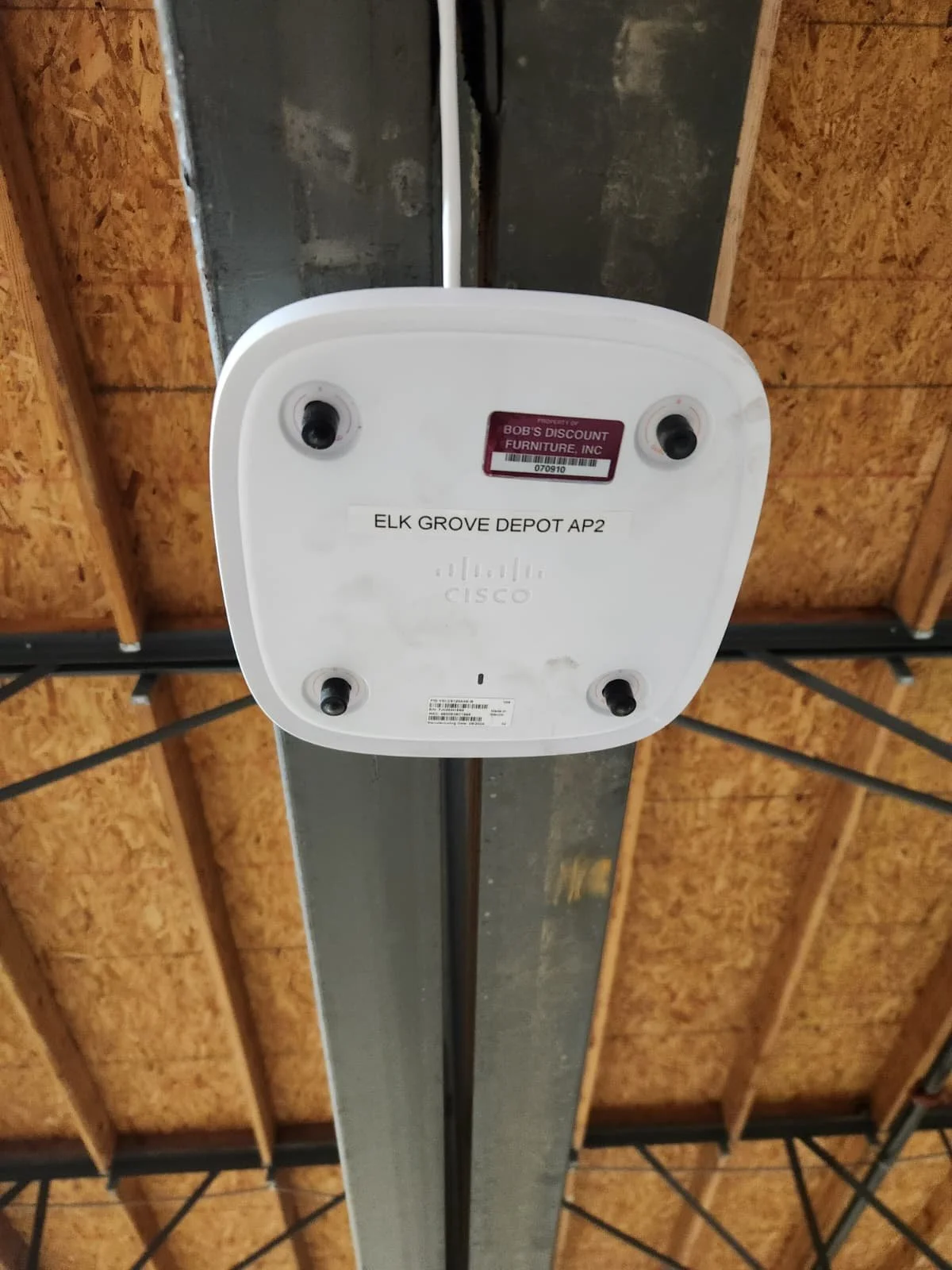 Ceiling-mounted observatory device with cables and a sticker, mounted on a metal beam in a warehouse or garage, with exposed wooden ceiling panels in the background.