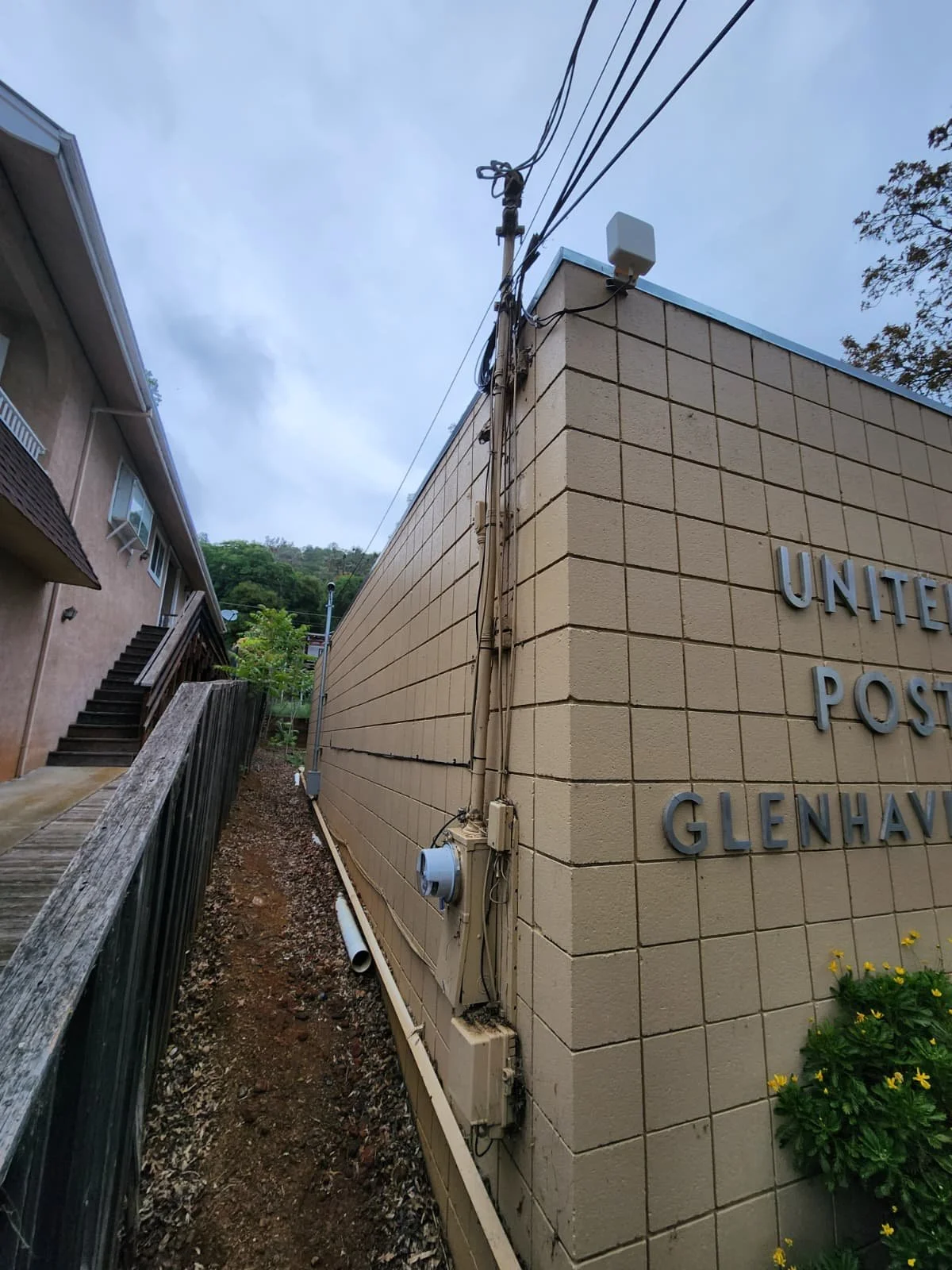 Side view of a beige brick United States Postal Service building with visible electrical wires and meters, with a wooden staircase and house on the left, and some greenery in the background.