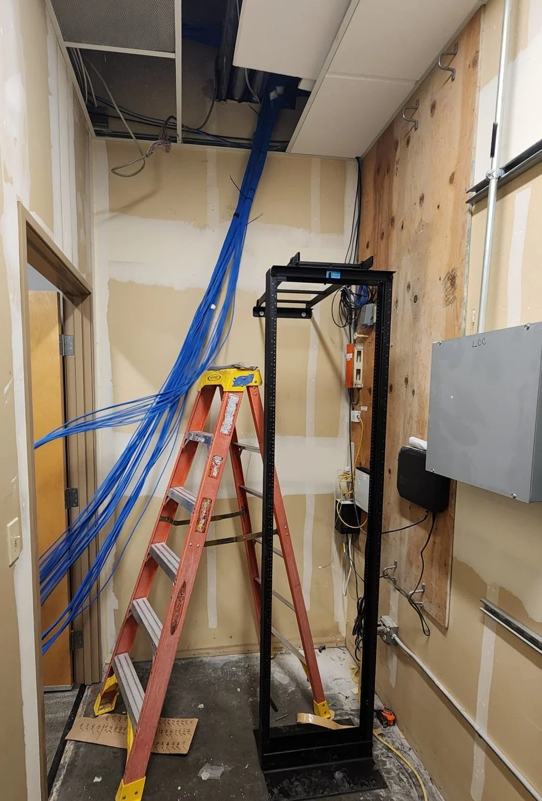 A small utility or server room under construction or renovation with drywall, blue network cables hanging from the ceiling, a red ladder, a black metal rack frame, and various electrical components and wires.