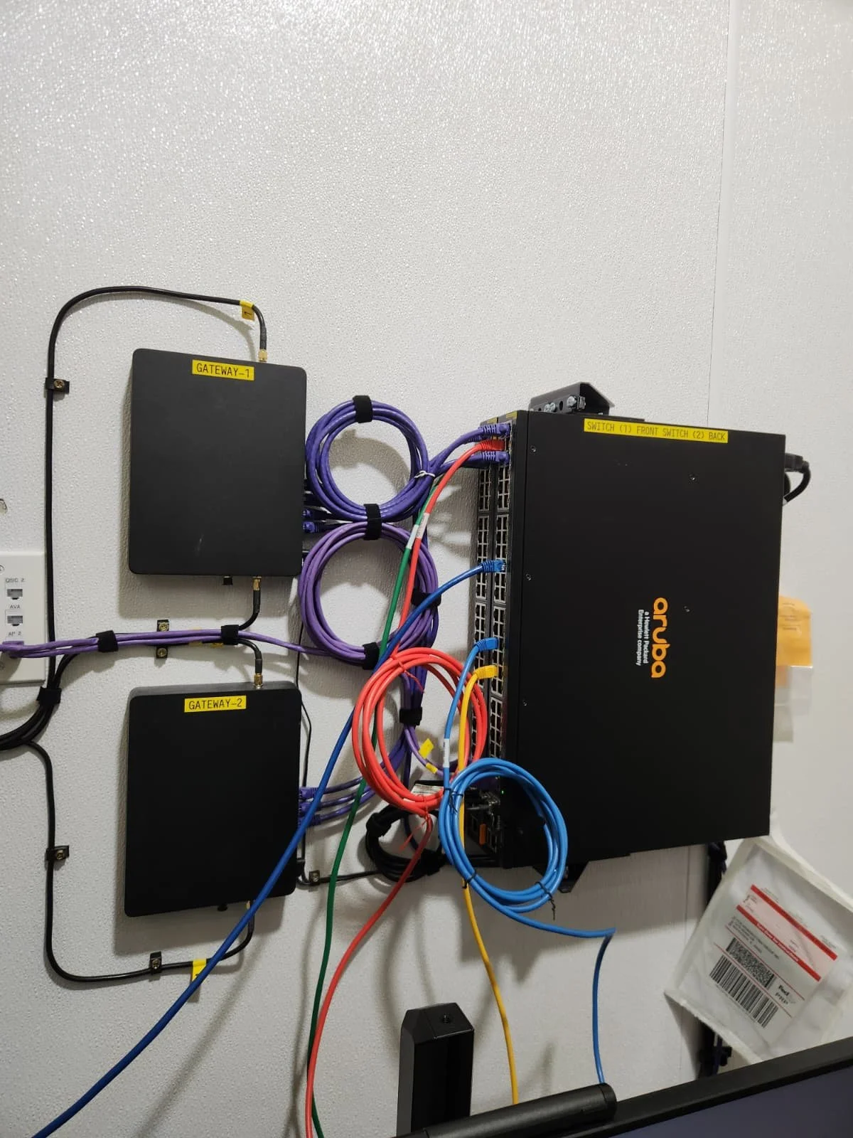 Close-up of a wall-mounted network and power equipment with several colorful cables connected to black labeled boxes, including a large black aruba switch, with labels 'GATEWAY-1', 'GATEWAY-2', and 'SWITCH (1) FRONT SWITCH (2) BACK'.