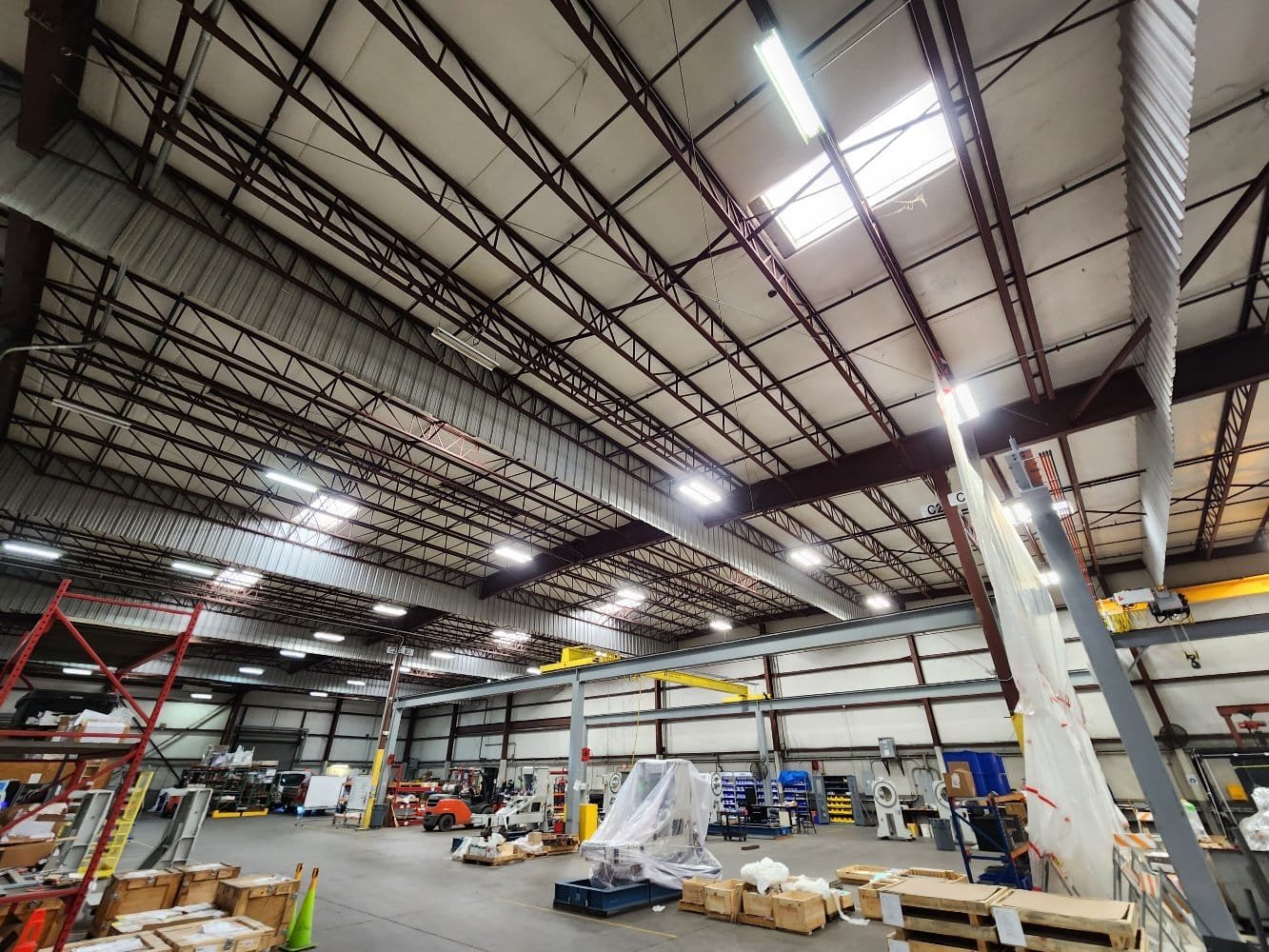 Interior of a warehouse with high ceilings, various equipment, crates, pallets, and shelves, some covered in plastic.