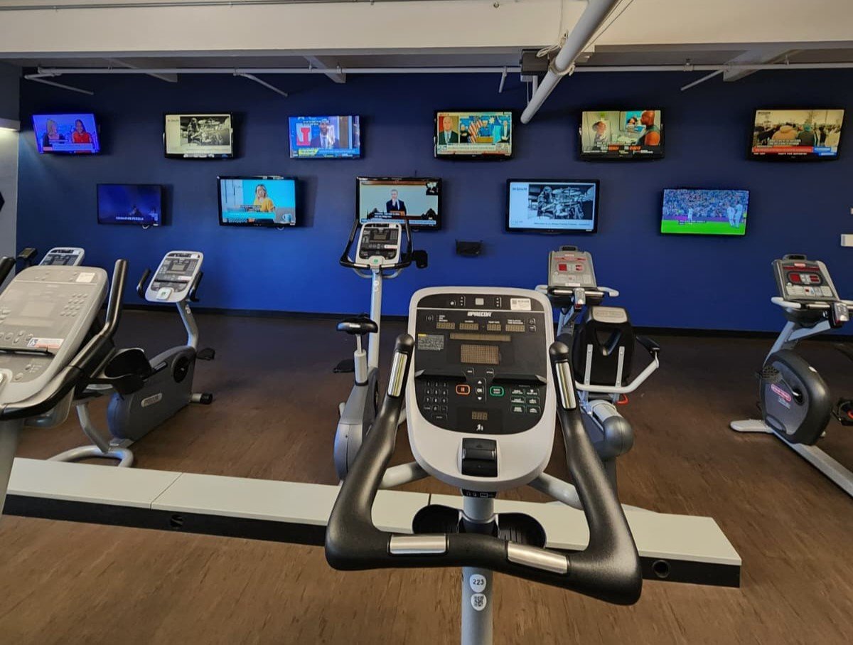 Interior of a gym with exercise equipment and multiple wall-mounted TVs.